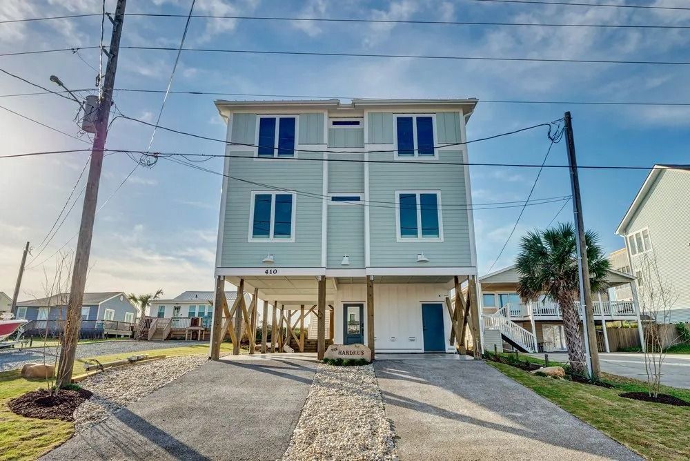 Three-story beach house on stilts with a driveway, blue-gray exterior, and utility poles.