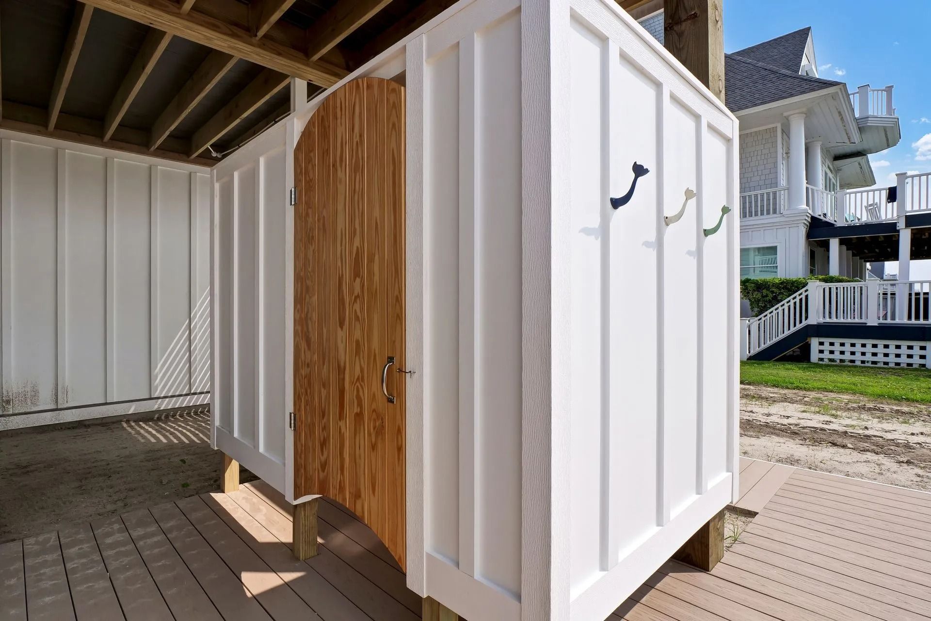 Outdoor shower with white paneling and wooden door, near a house with stairs.