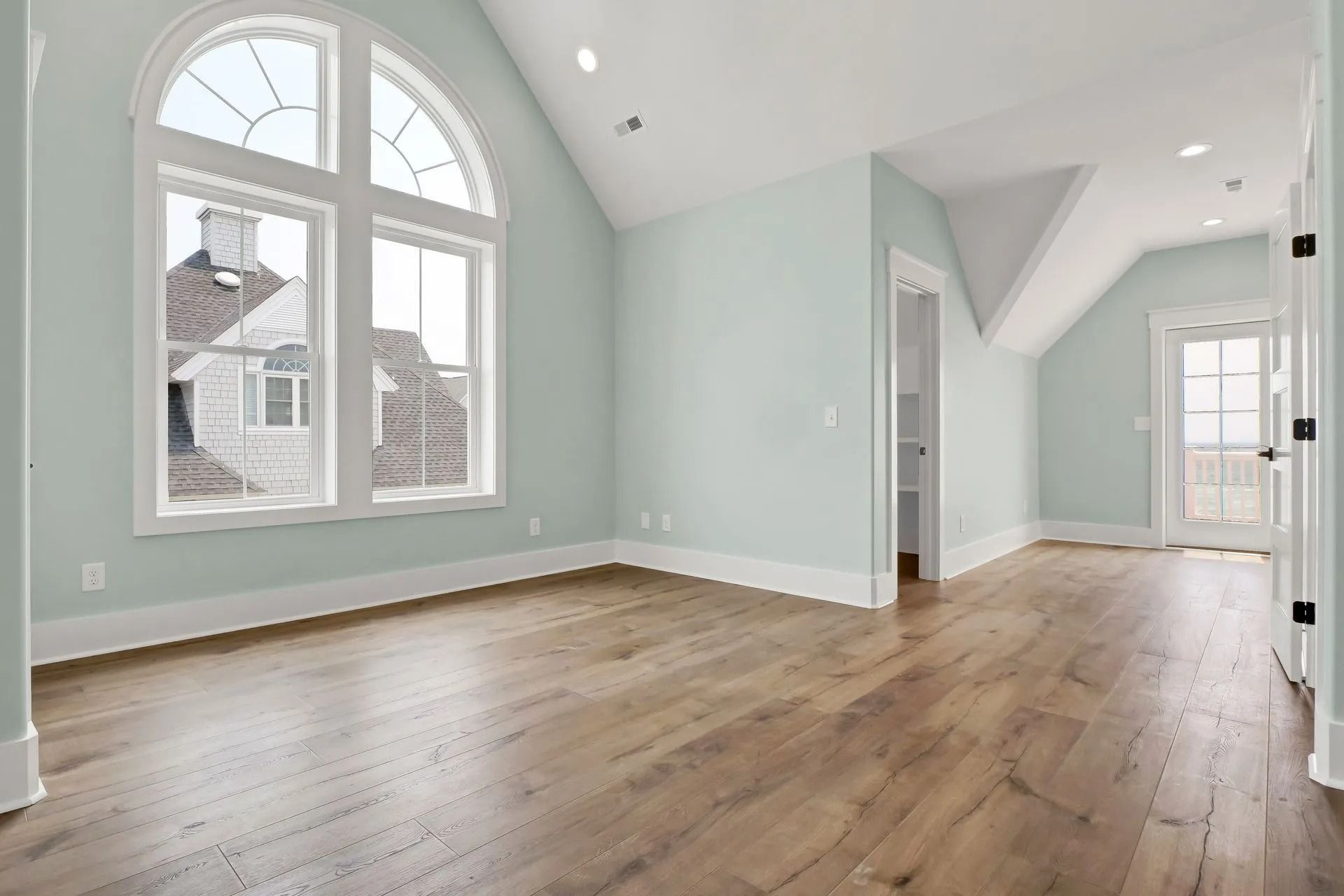 Empty room with hardwood floors, arched window, light blue walls, and a white trim.