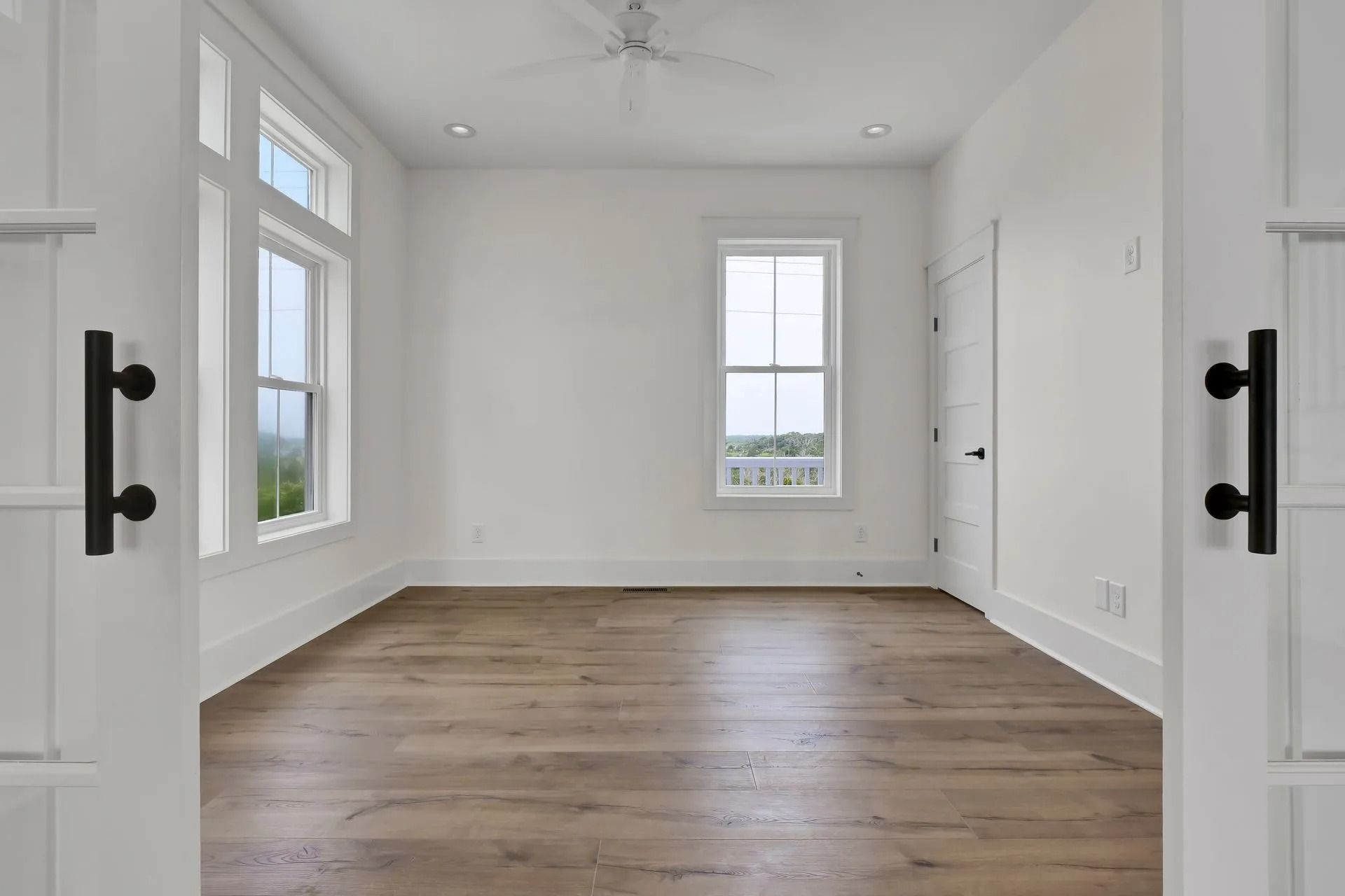 Empty white room with light wood floors and windows. Black door handles and trim.