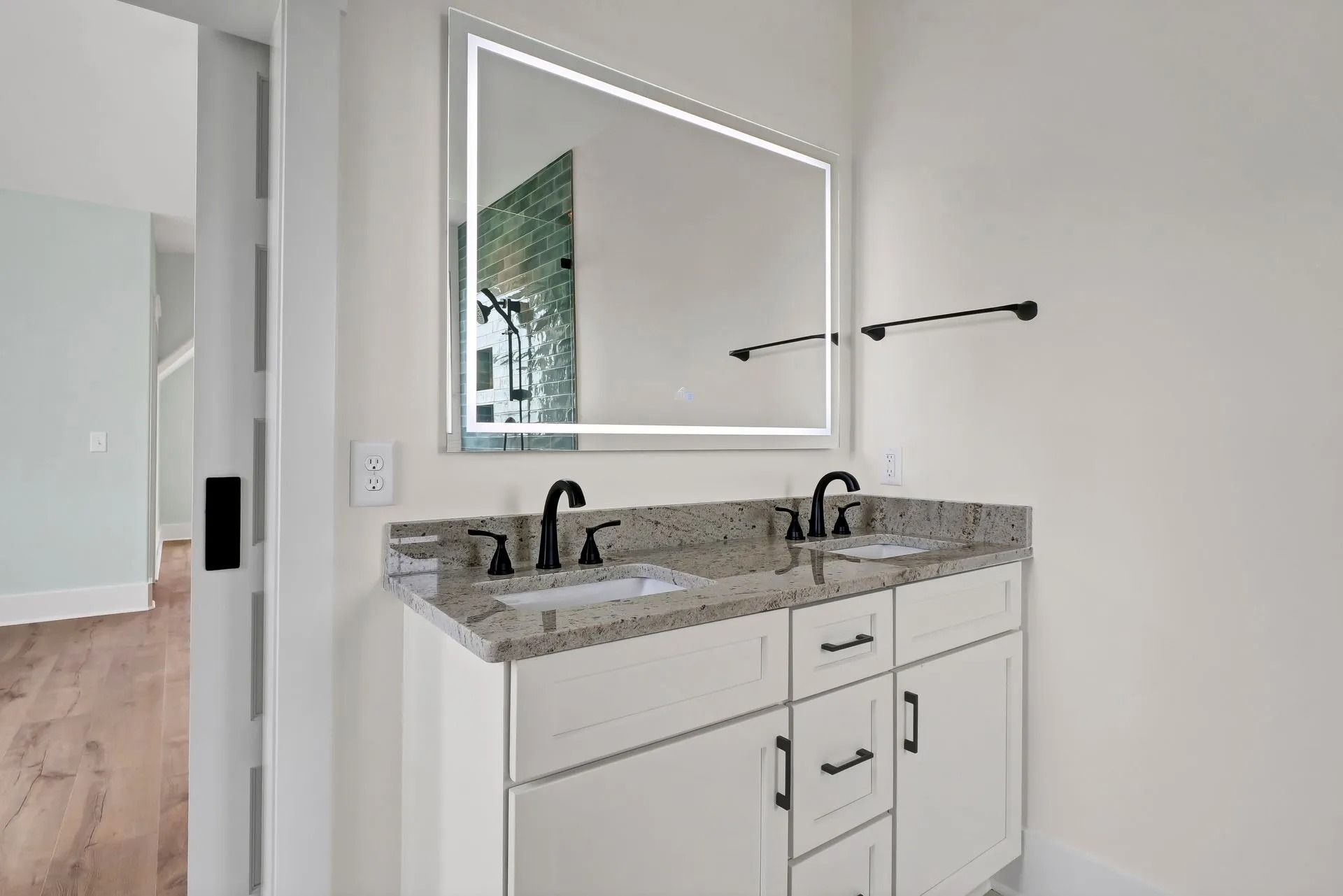 Modern bathroom with white double vanity, granite countertop, lit mirror, and black fixtures.