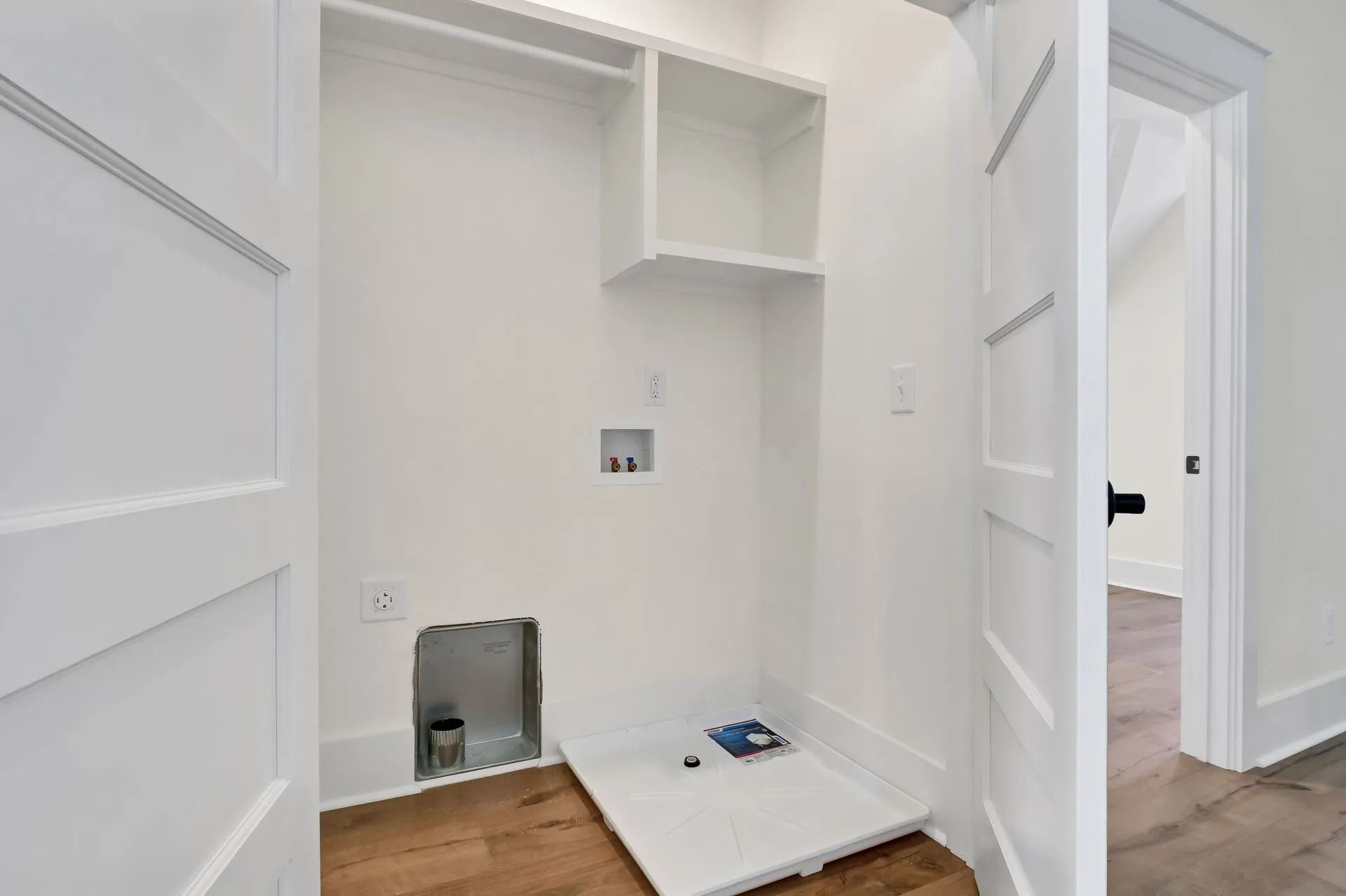 Empty white laundry room with open doorway, shelving, and water hookups.