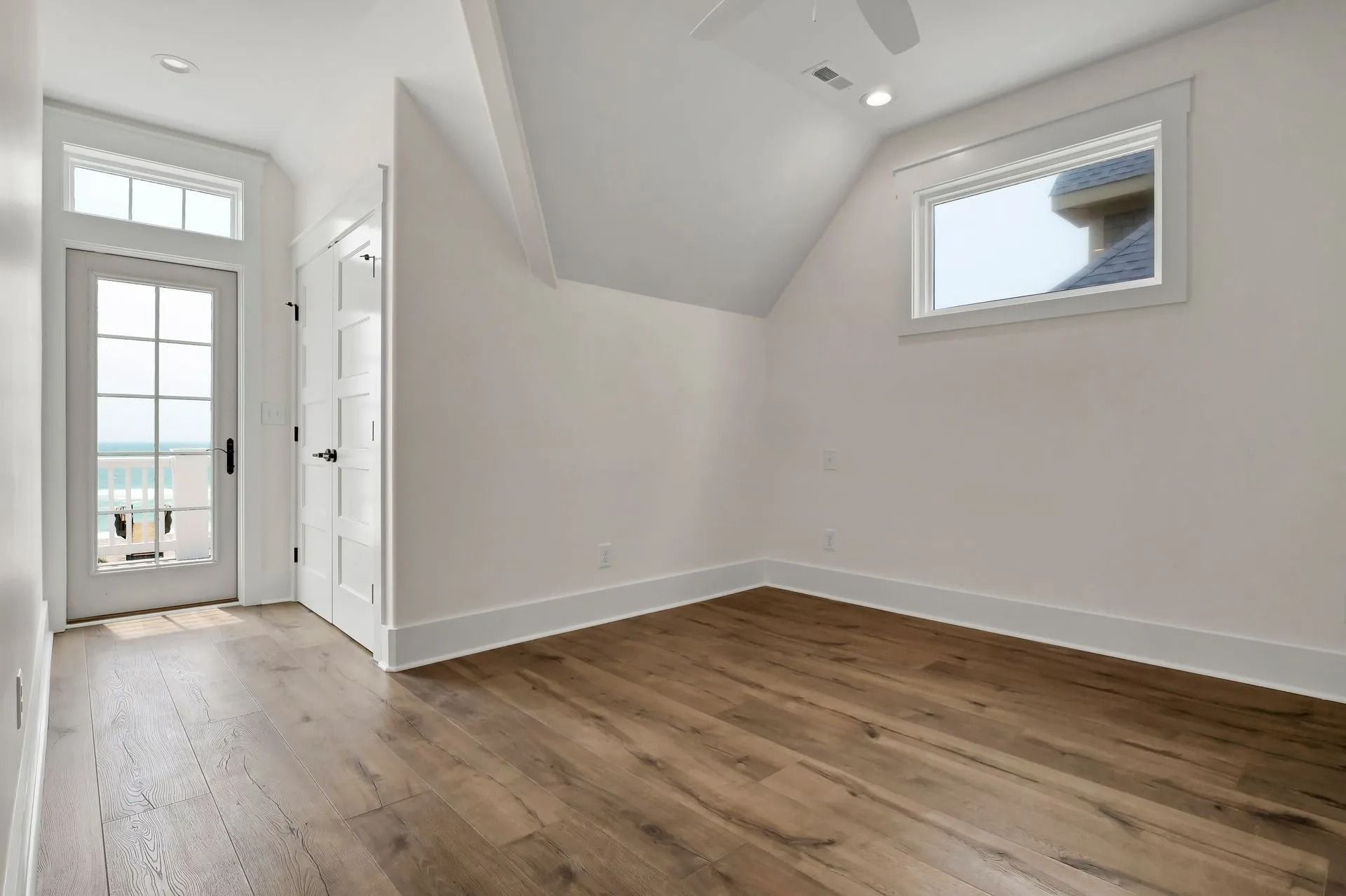 Empty room with hardwood floors, white walls, and a door leading to a balcony with a beach view.