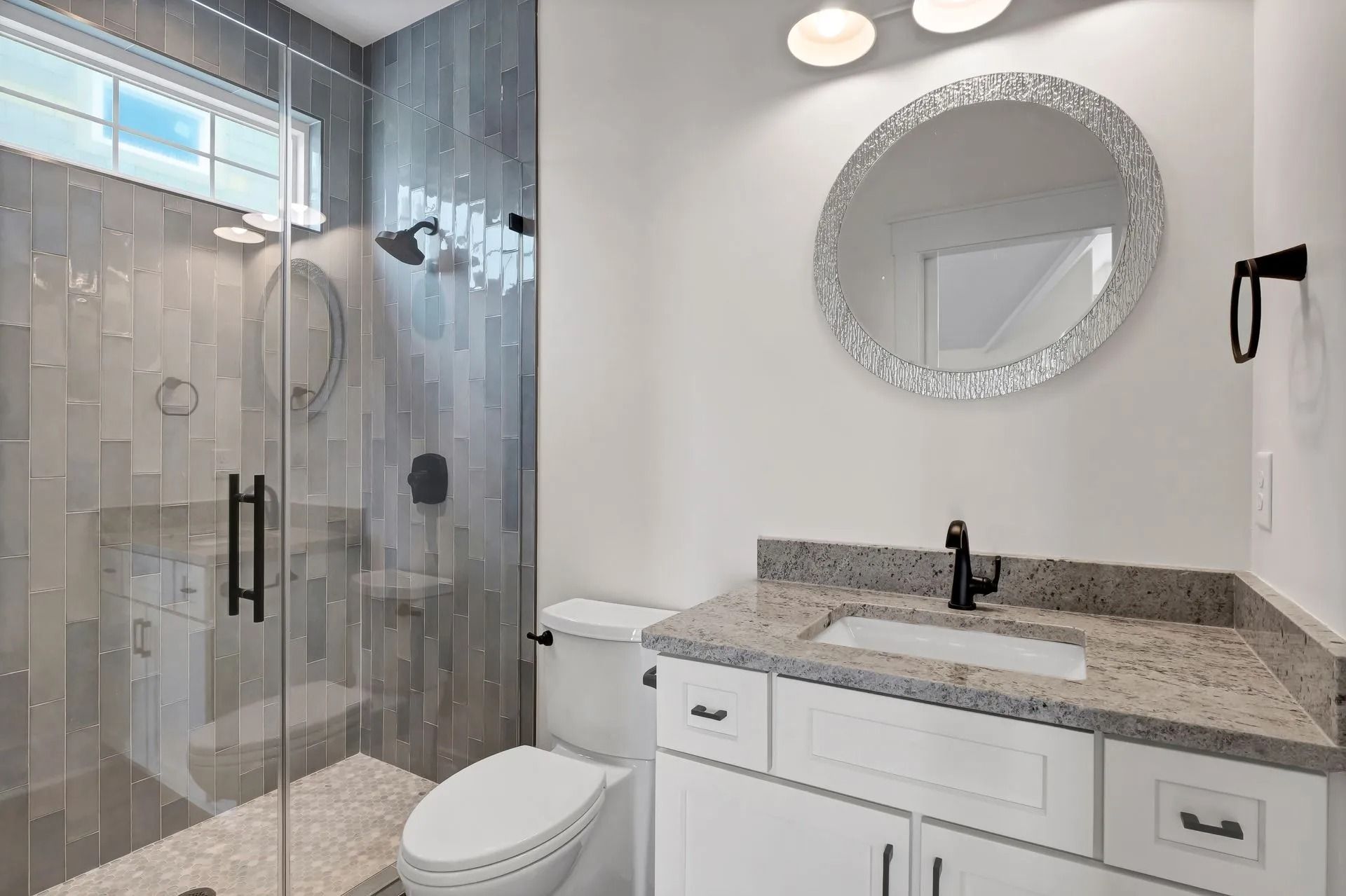 Bathroom with a glass shower, white vanity, round mirror, and gray tile walls.