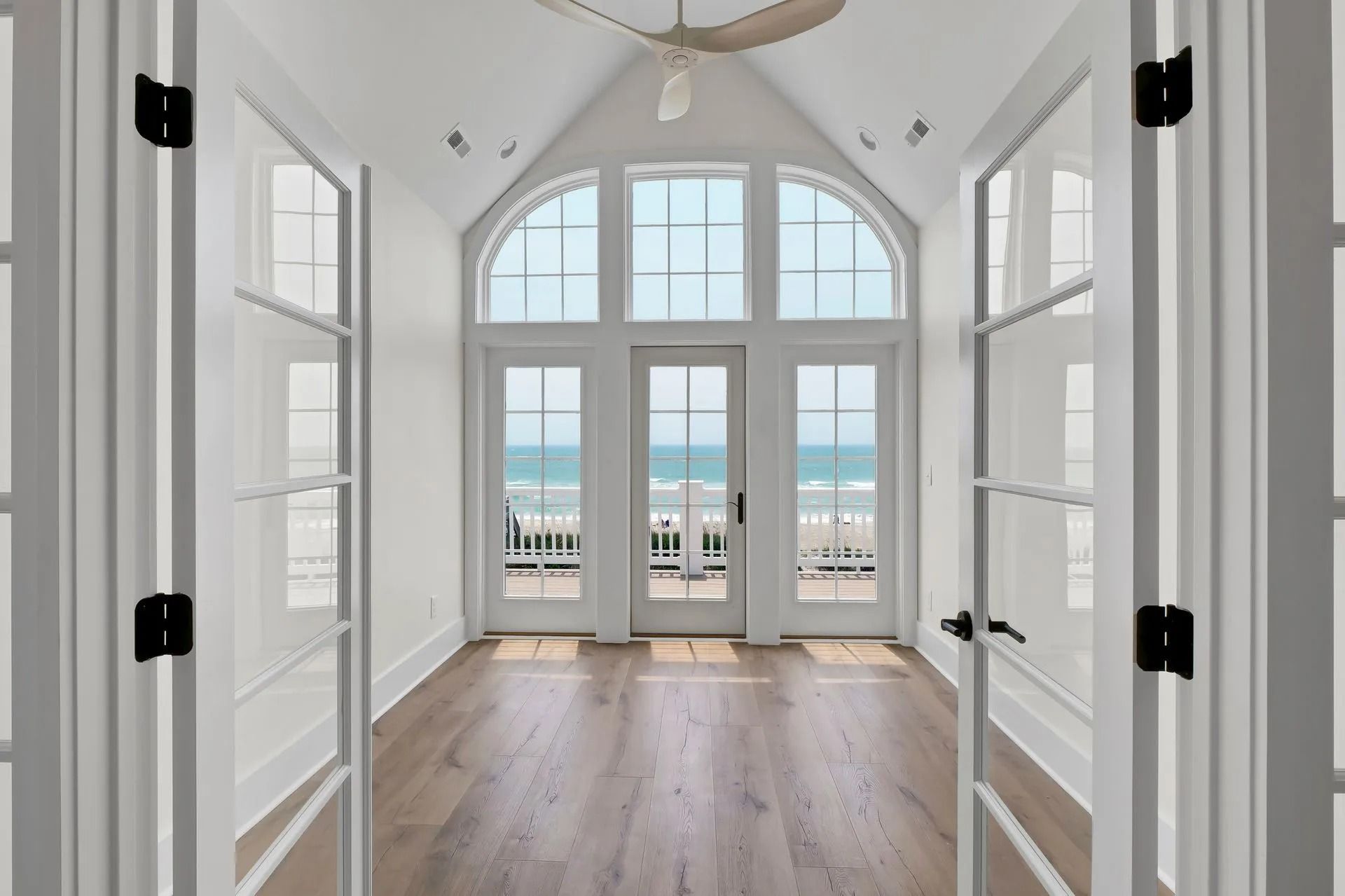 Bright room with ocean view, arched window doors, wood floors, white walls, and open white doors.