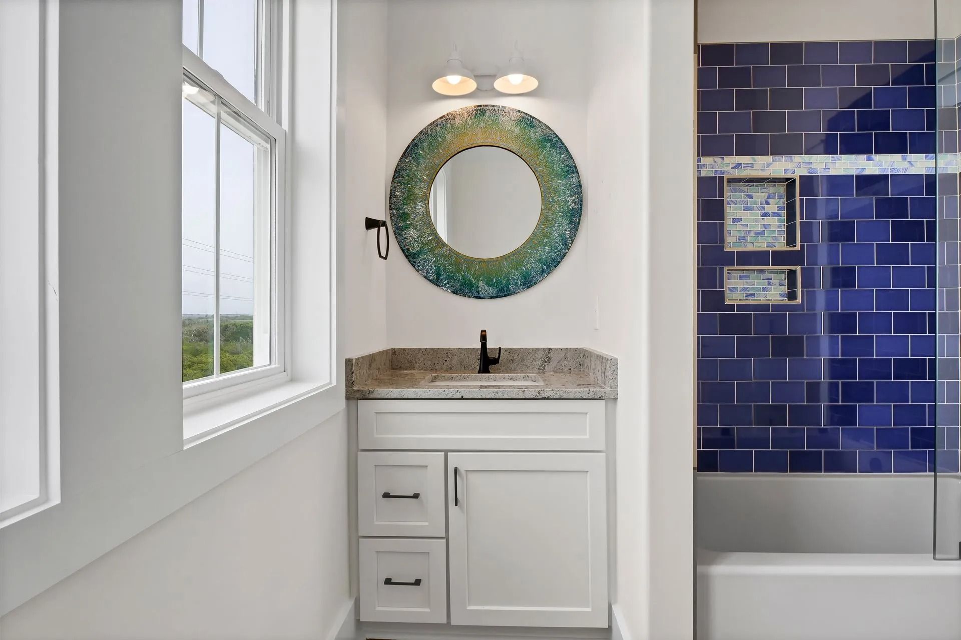 Bathroom with white vanity, round mirror, blue tile shower, and a window.
