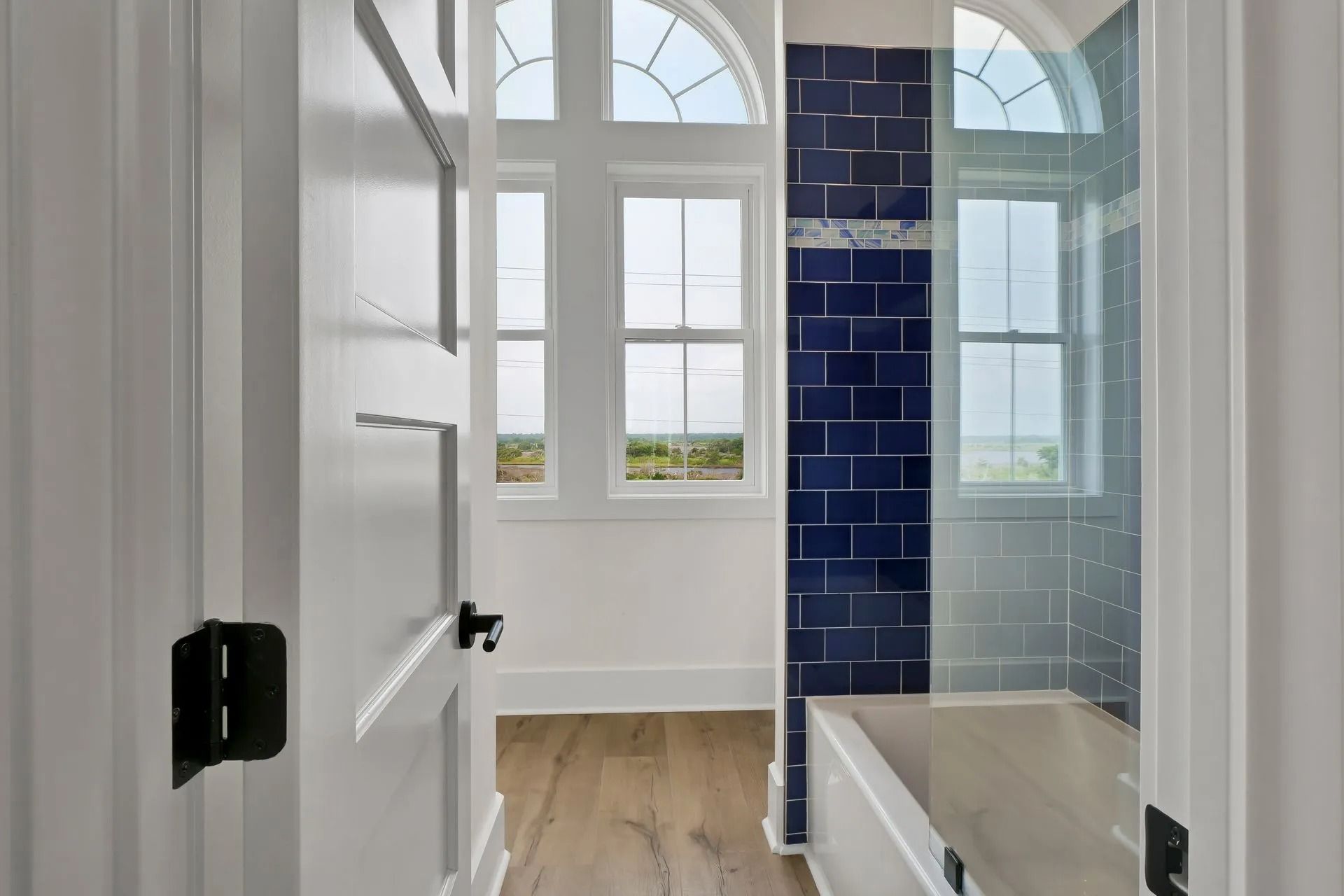Bathroom entryway with white door, wood floor, blue tiled shower, and arched windows.