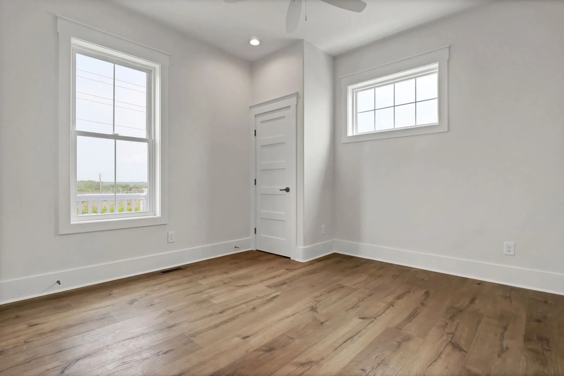 Empty room with hardwood floors, white walls, two windows, and a closed door.