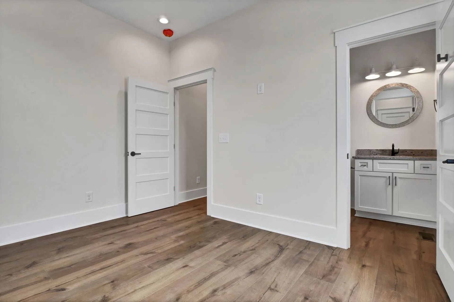 Empty room with hardwood floors, white walls, and a doorway leading to a bathroom.