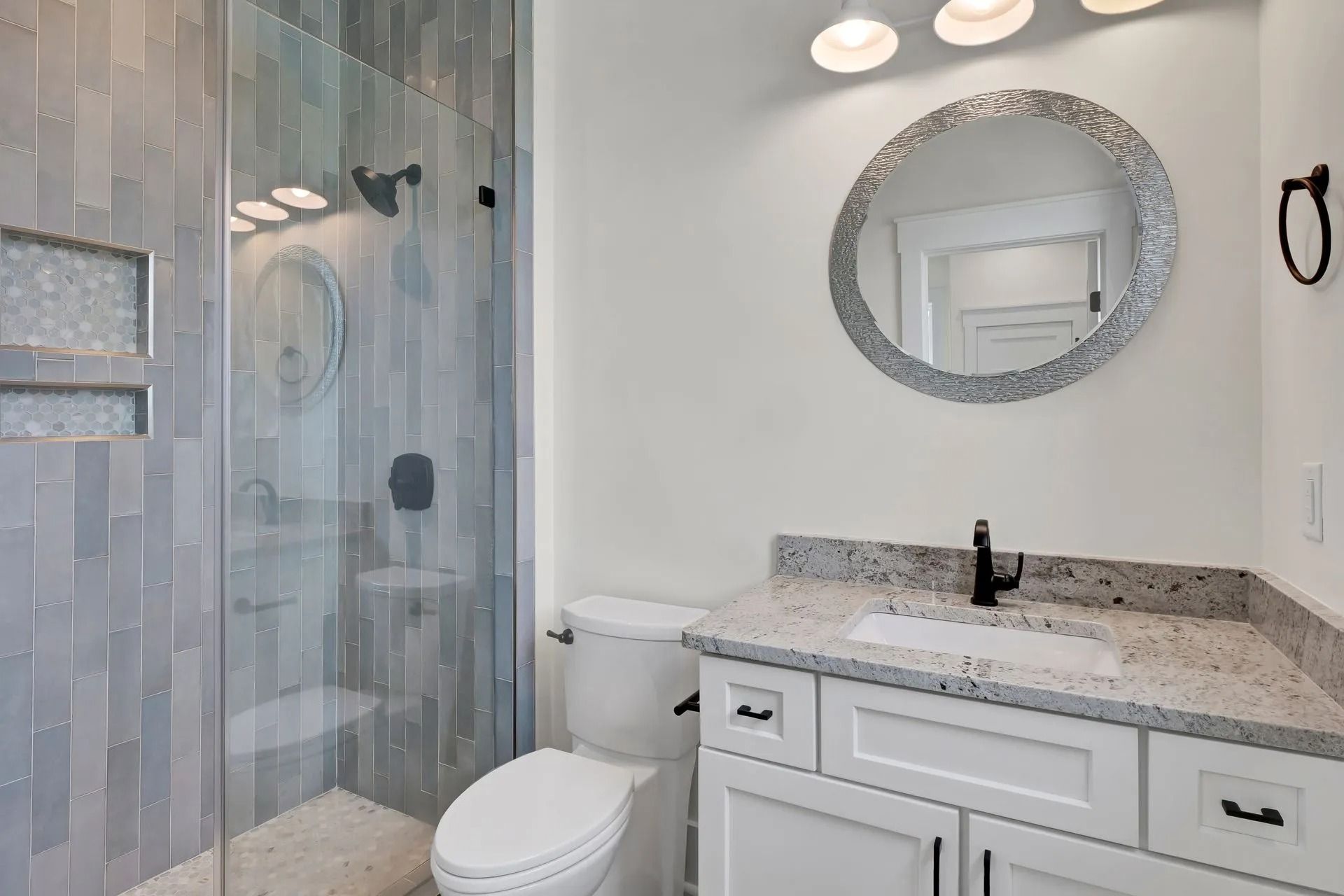 Bathroom with shower, toilet, white vanity with speckled countertop, and round mirror.