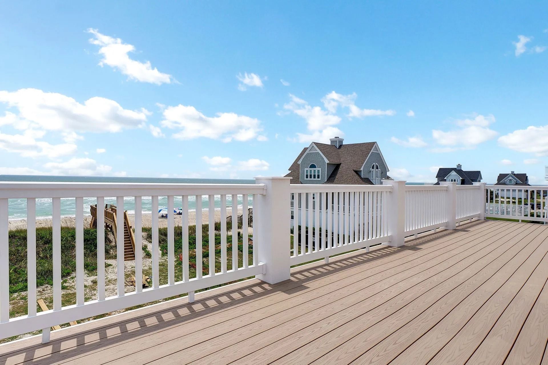 Wooden deck with white railing overlooking a beach and houses under a blue sky with clouds.