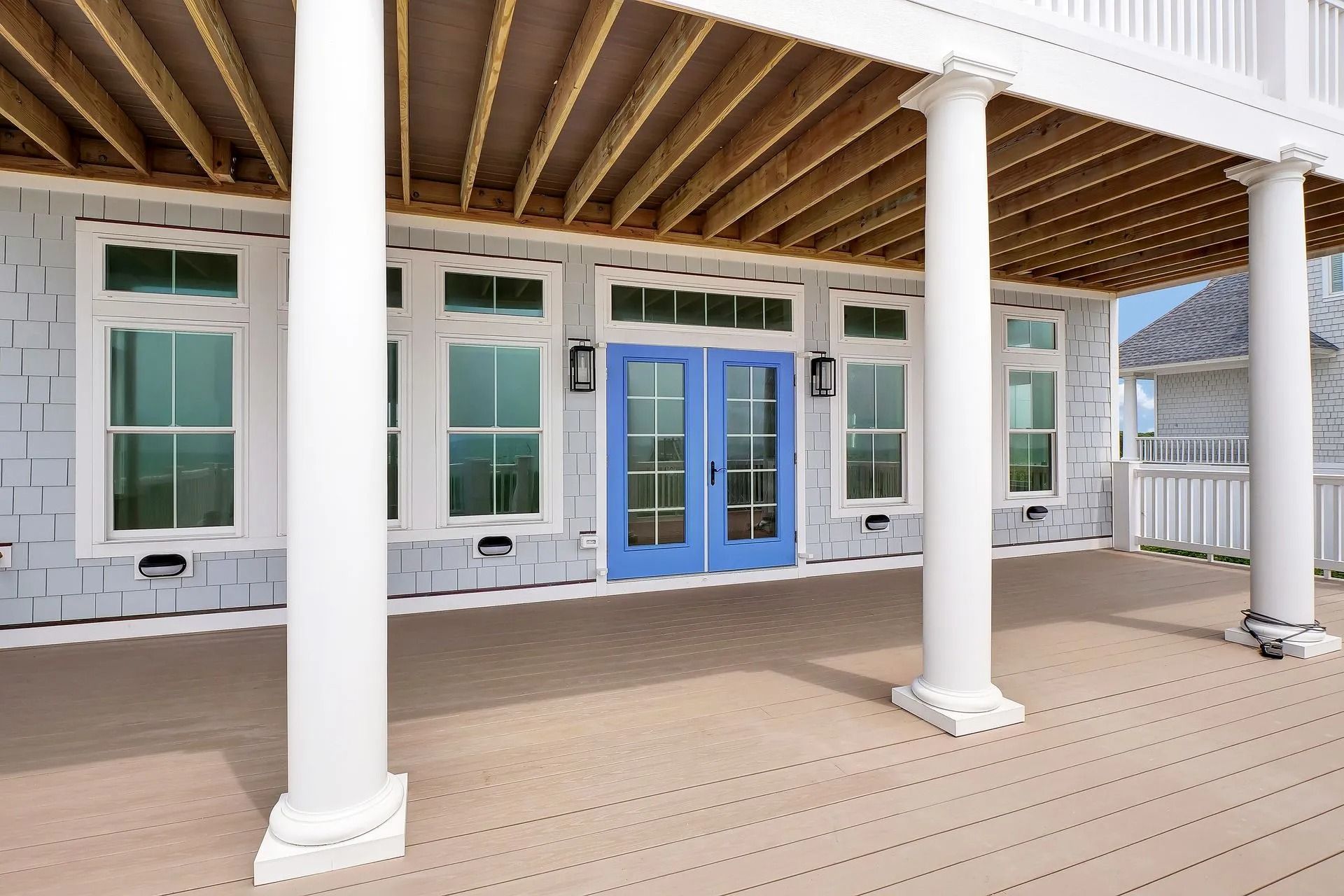 A porch with white columns, blue double doors, and windows, beneath a deck with wooden planks.