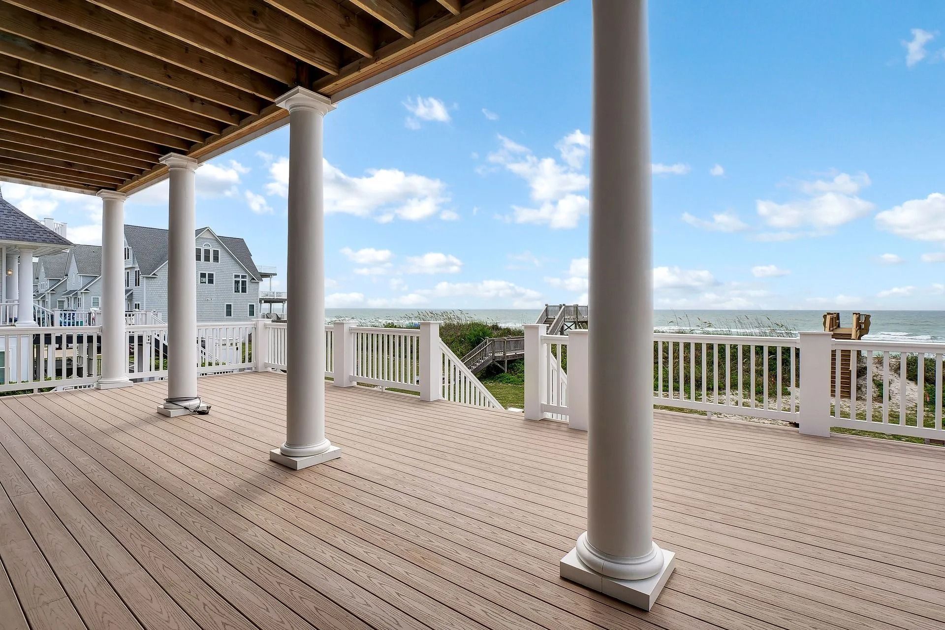 Wooden deck with white columns overlooking ocean. Blue sky with clouds.