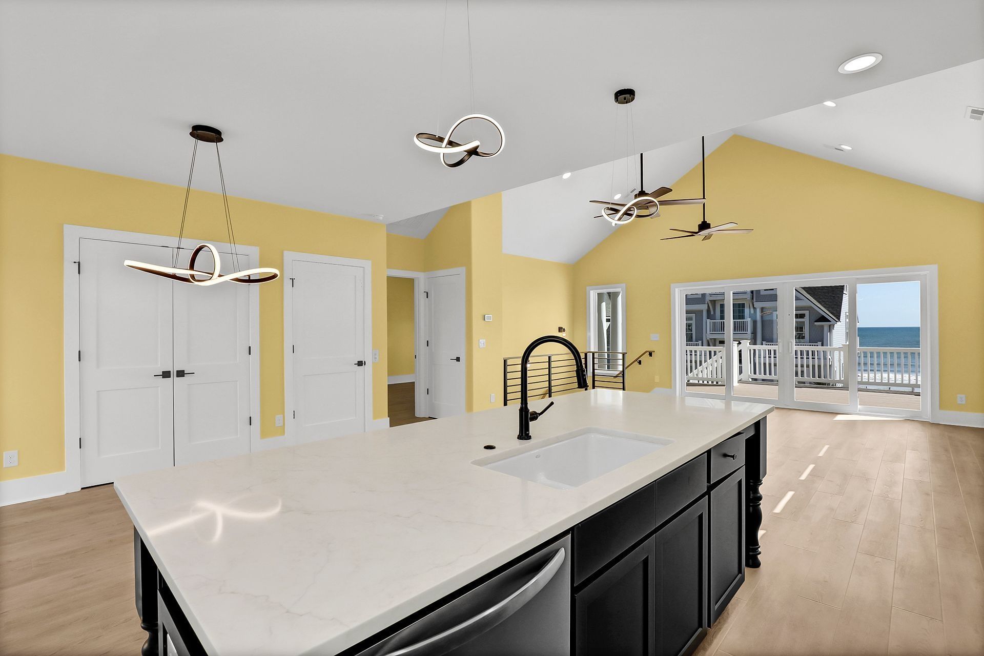Modern kitchen with white island, black cabinets, and yellow walls, overlooking a deck and ocean view.
