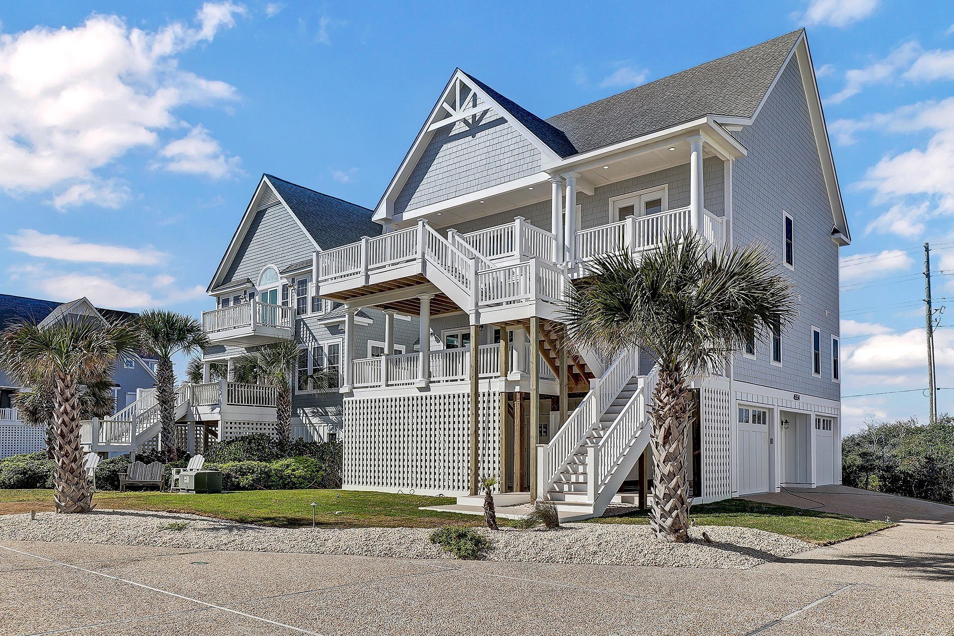 Beach house with light blue siding, white trim, and a gray roof, set on a sandy lot under a partly cloudy sky.