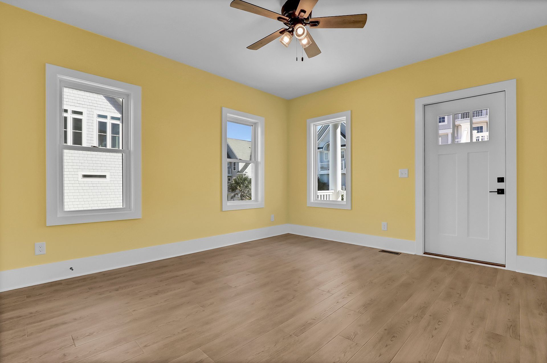 Empty room with yellow walls, white trim, wood floors, and ceiling fan. White door and windows.