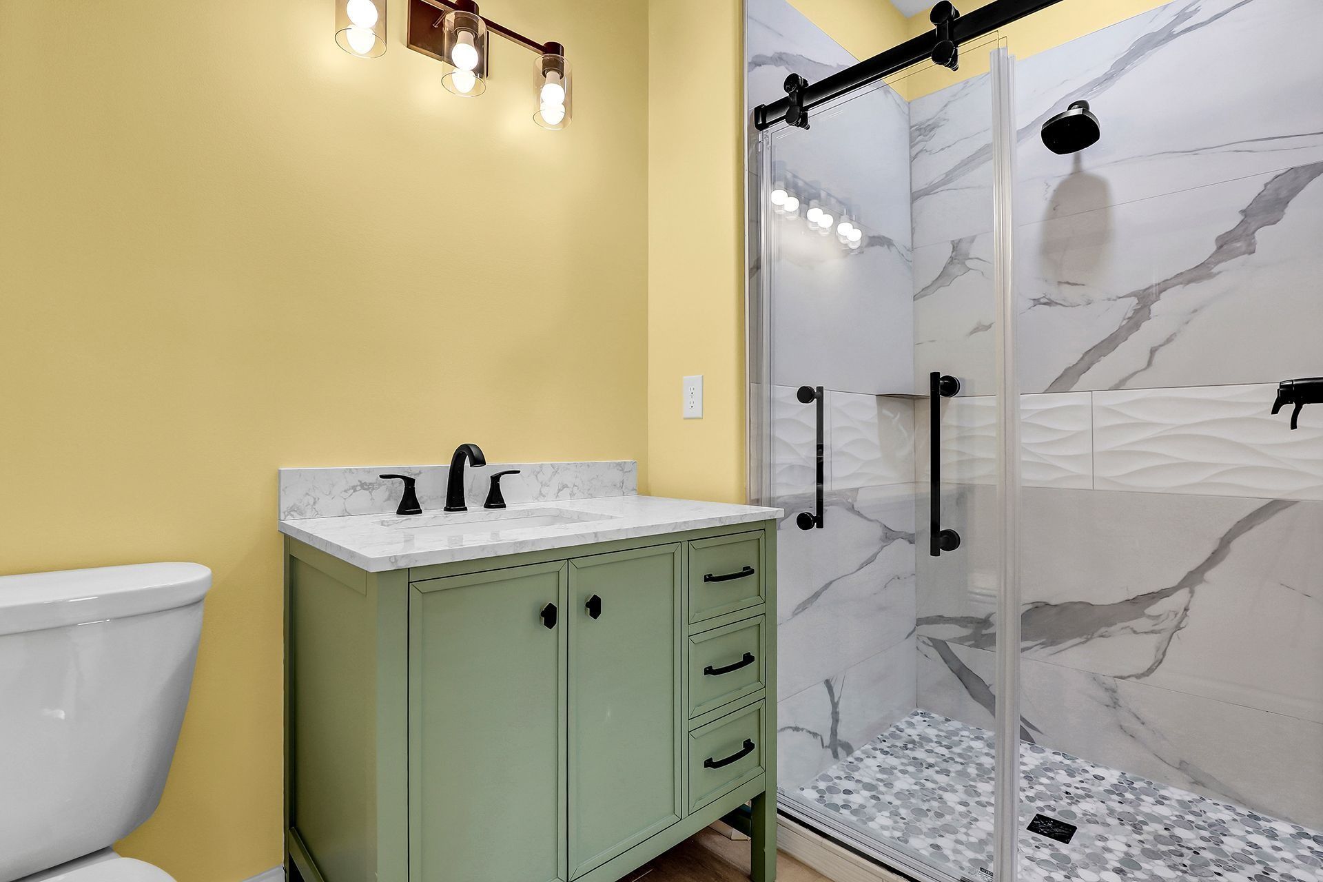 Bathroom with a light green vanity, white countertop, and glass shower with marble-like tile. Yellow wall and black hardware.