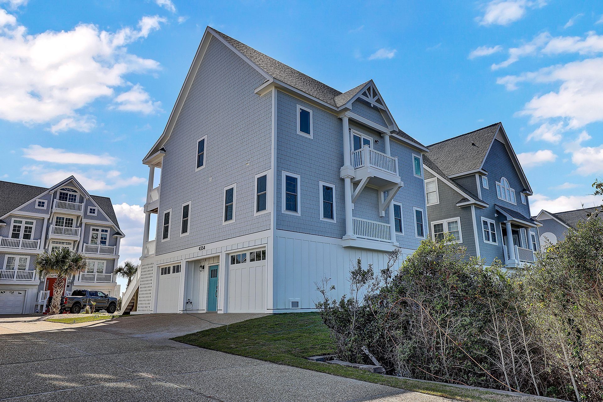 Blue and white coastal house with balconies, garages, and gravel driveway under a partly cloudy sky.