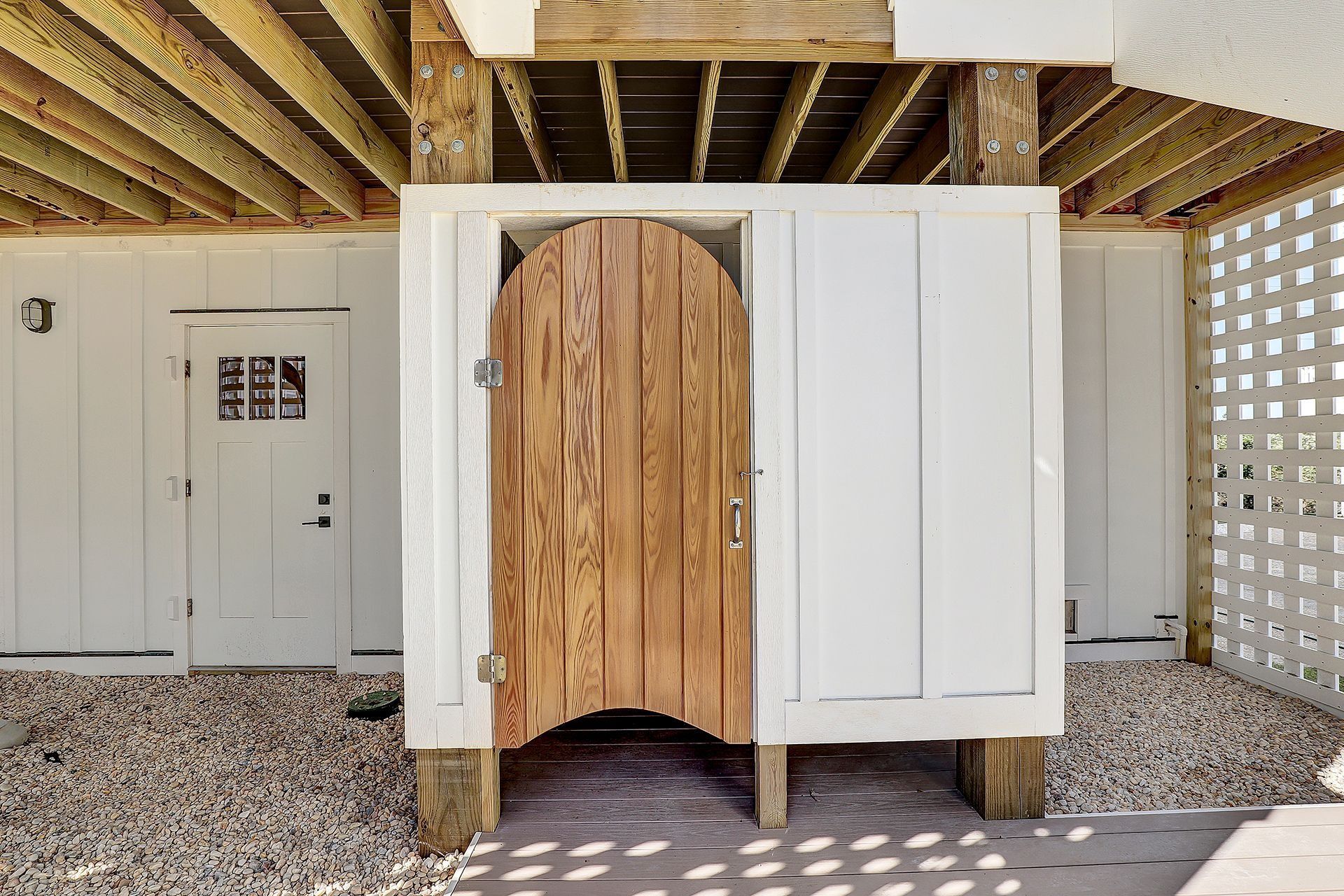 Outdoor shower with wooden door, set under a house on gravel. White siding and lattice with wooden beams.