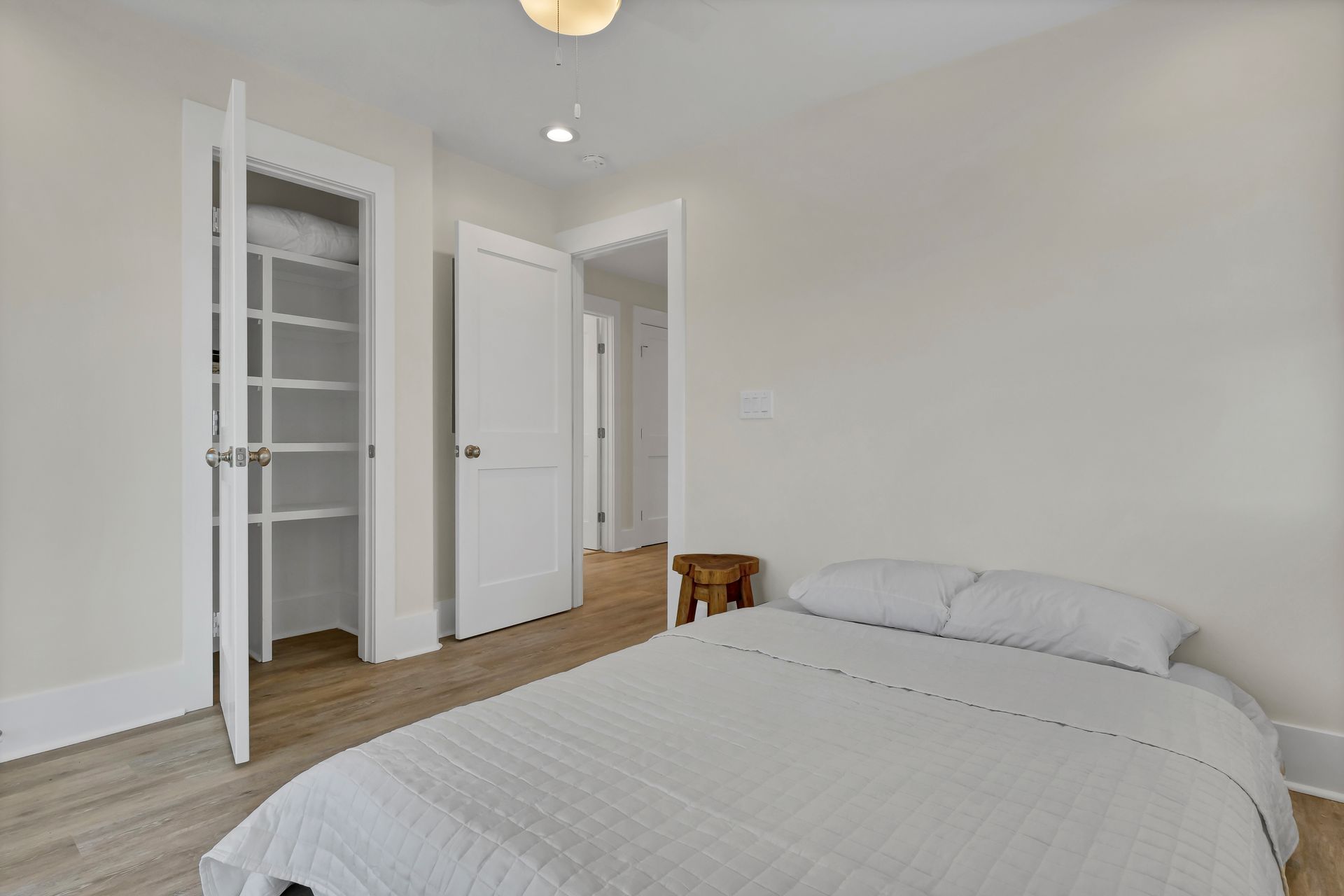 Bedroom with bed, open closet, door, and wooden stool. Light-colored walls and floor.