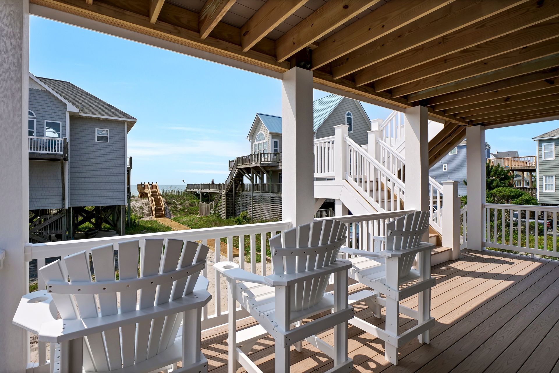 A covered porch with white Adirondack chairs overlooking beach houses and the ocean on a sunny day.