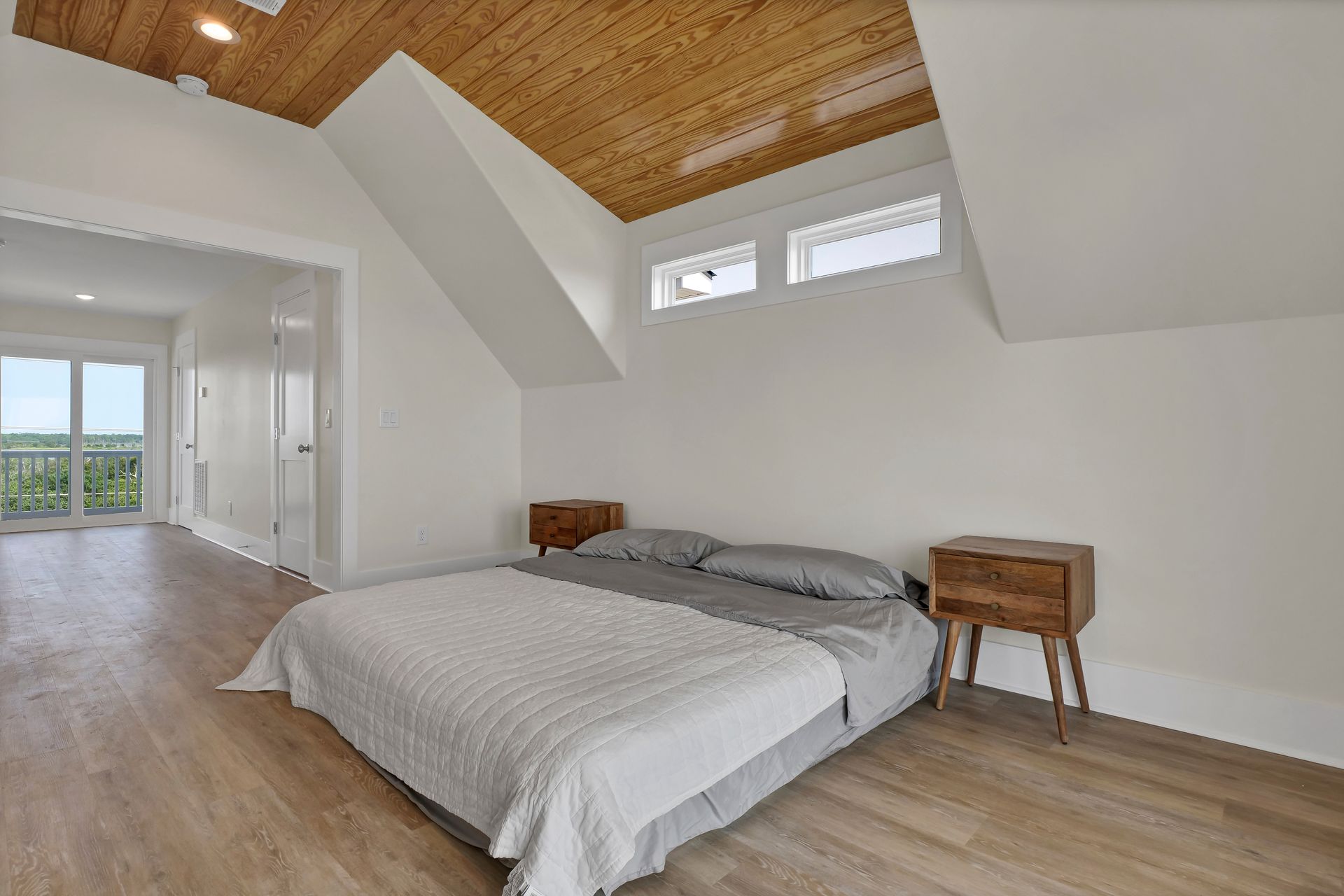 Bedroom with wood-paneled ceiling, bed with gray linens, two nightstands, and a door leading to a balcony.
