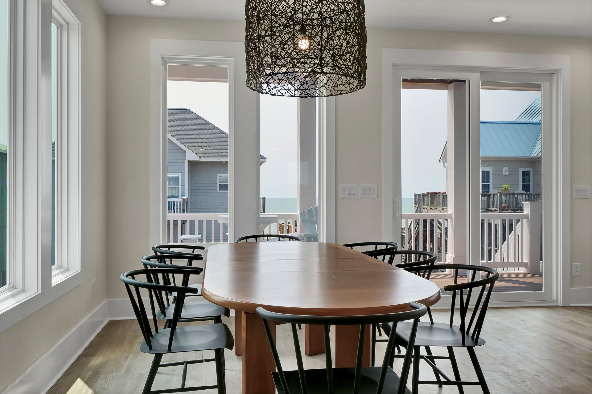 Dining room with a long wooden table, black chairs, and a decorative light fixture; windows with a view.