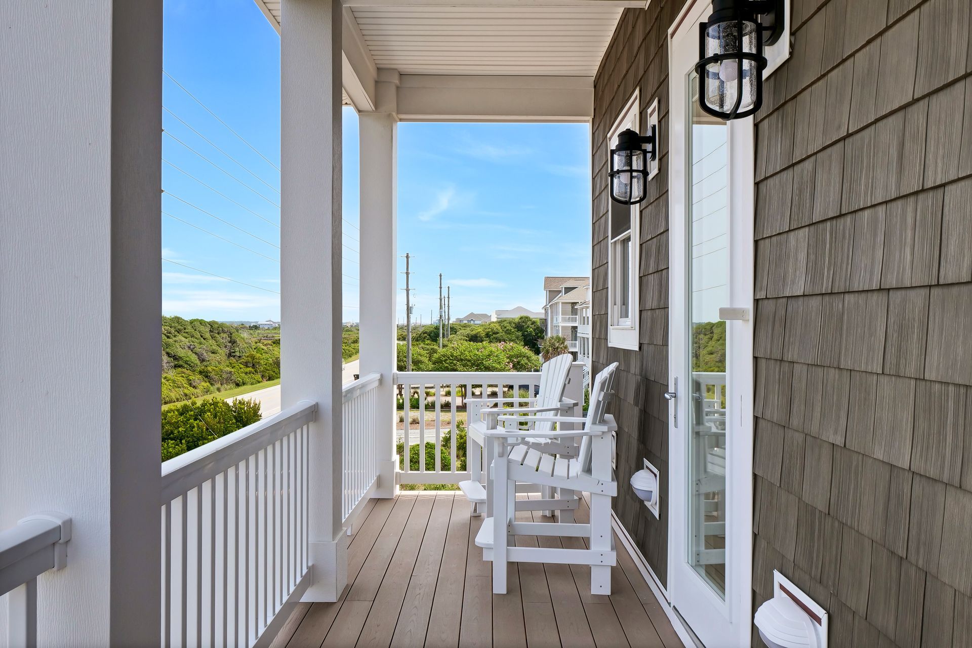 Wooden porch with white railings and chairs, overlooking green trees and a blue sky.