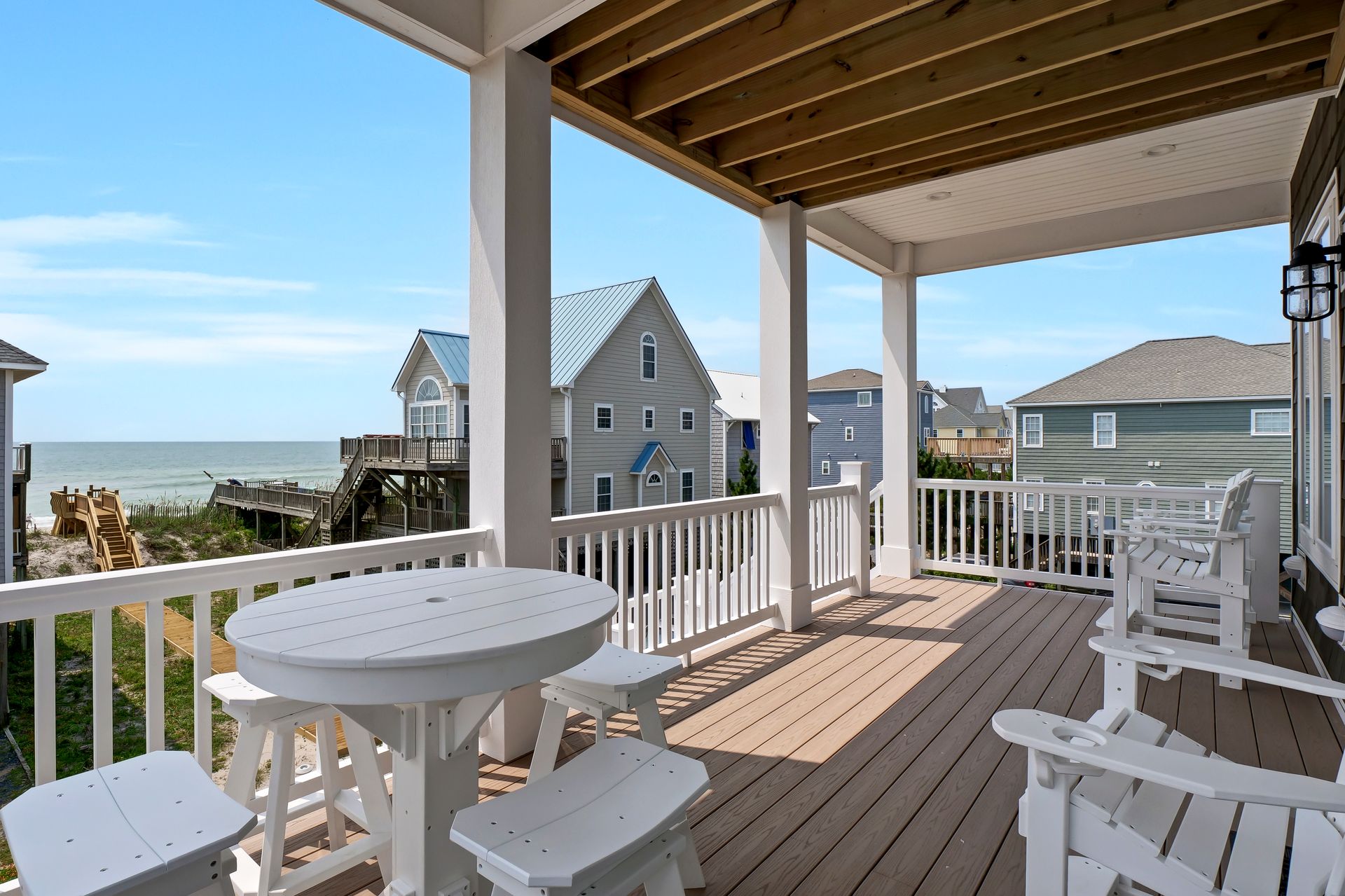 Covered deck with white furniture, overlooking ocean and neighboring houses on a sunny day.