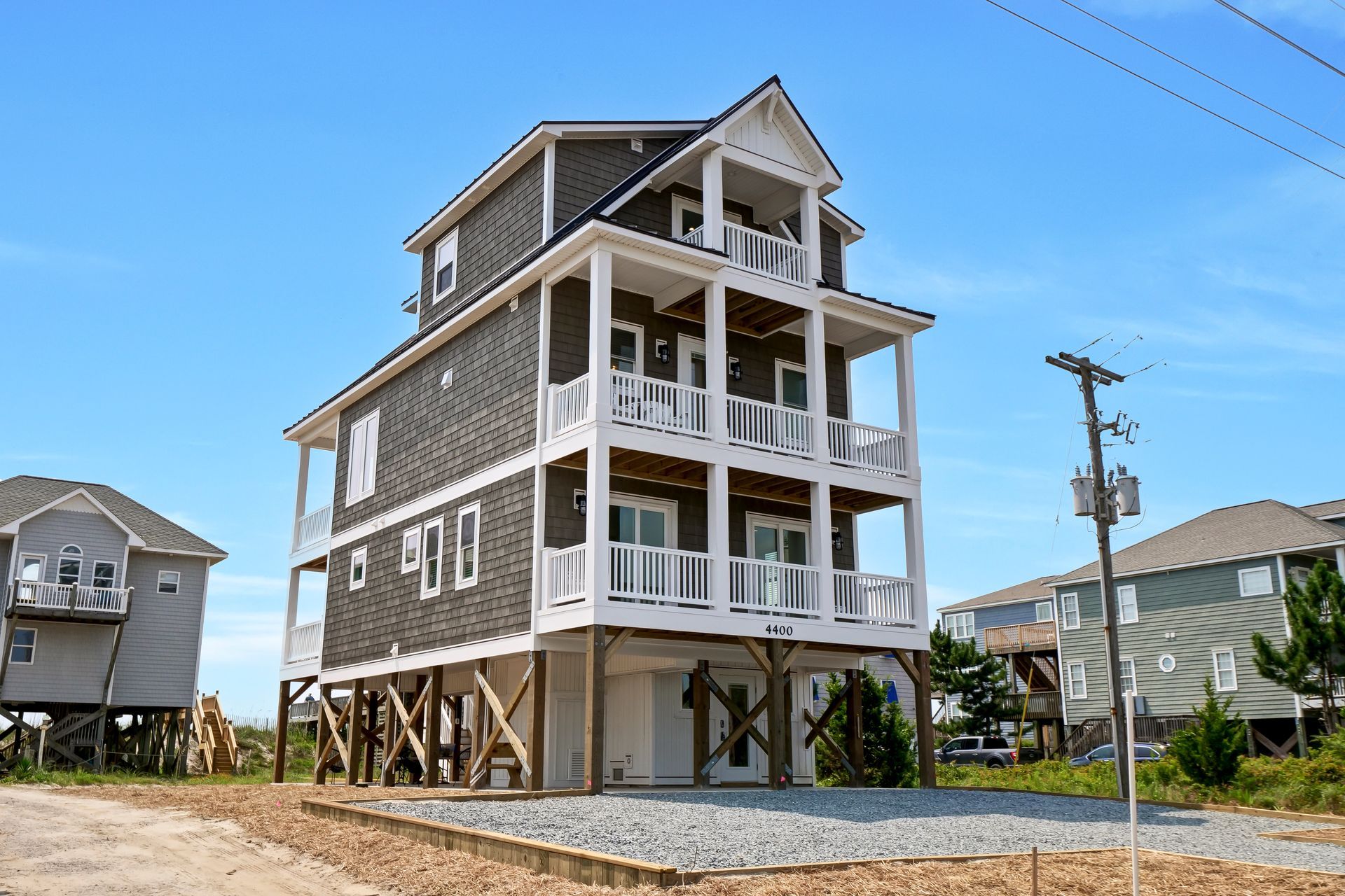 Three-story beach house on stilts with white railings, gray siding, and a blue sky.