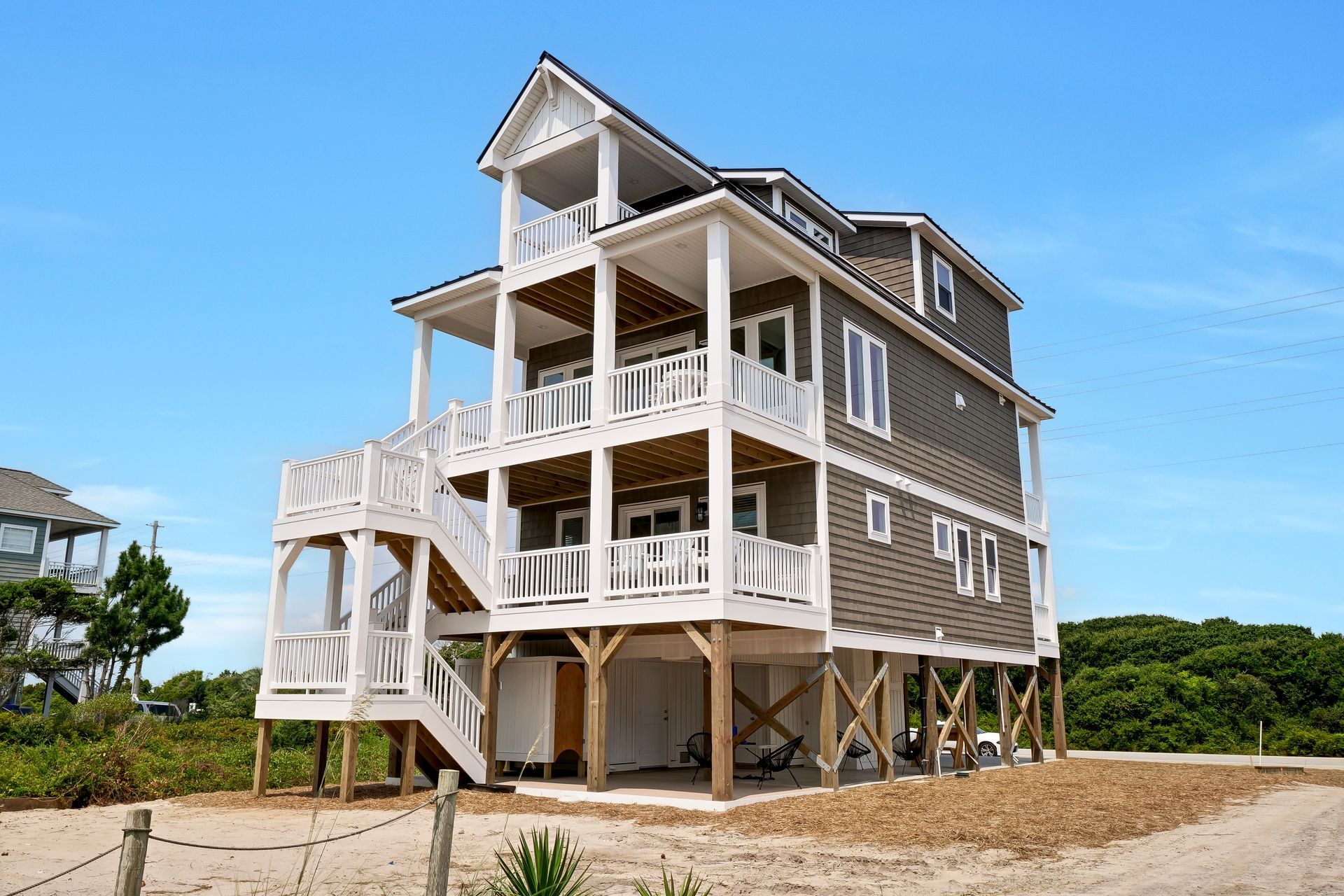 Three-story gray beach house on stilts with white railings and balconies, blue sky backdrop.