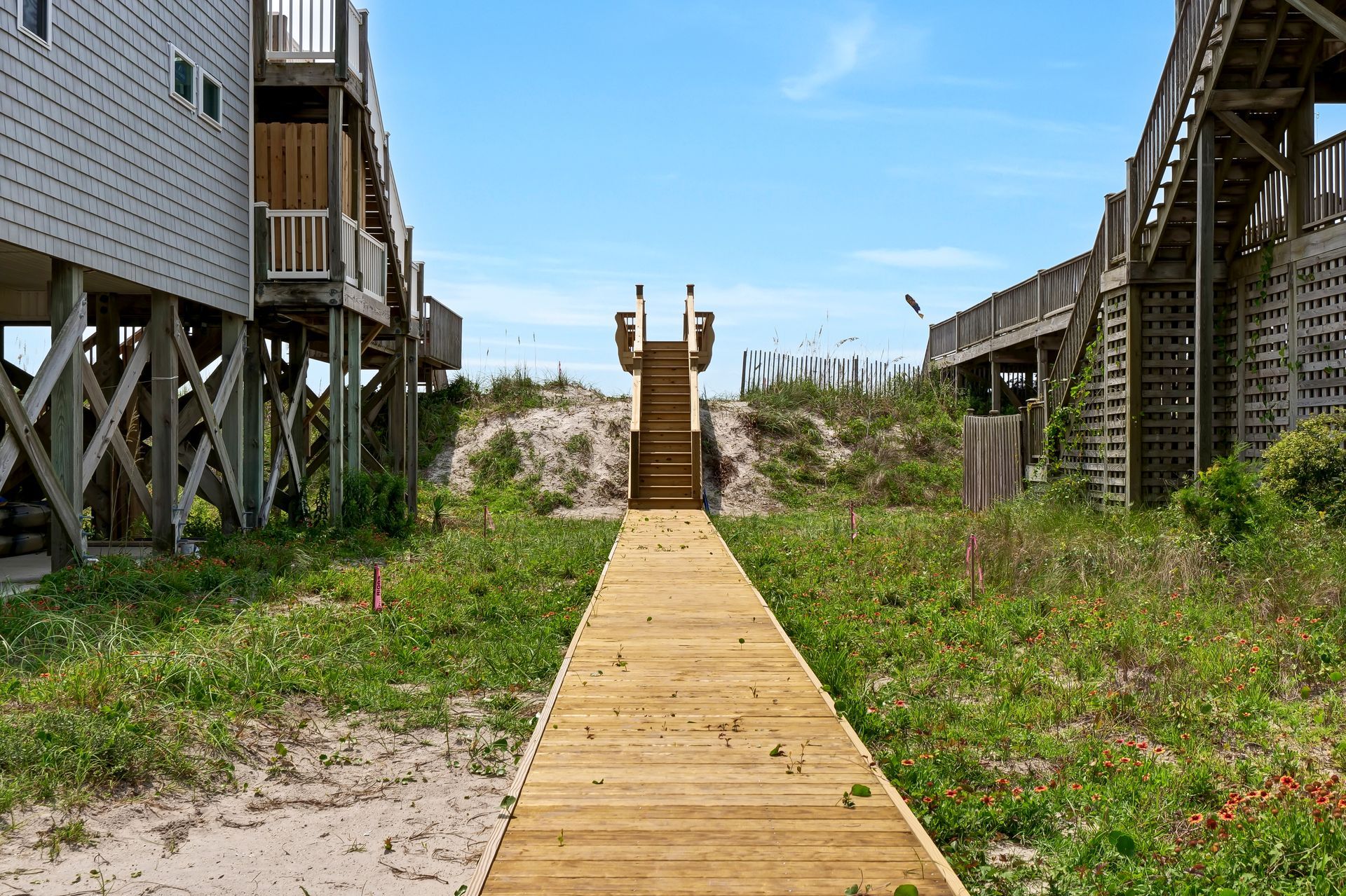 Wooden boardwalk leading to beach stairs, flanked by buildings, under blue sky.