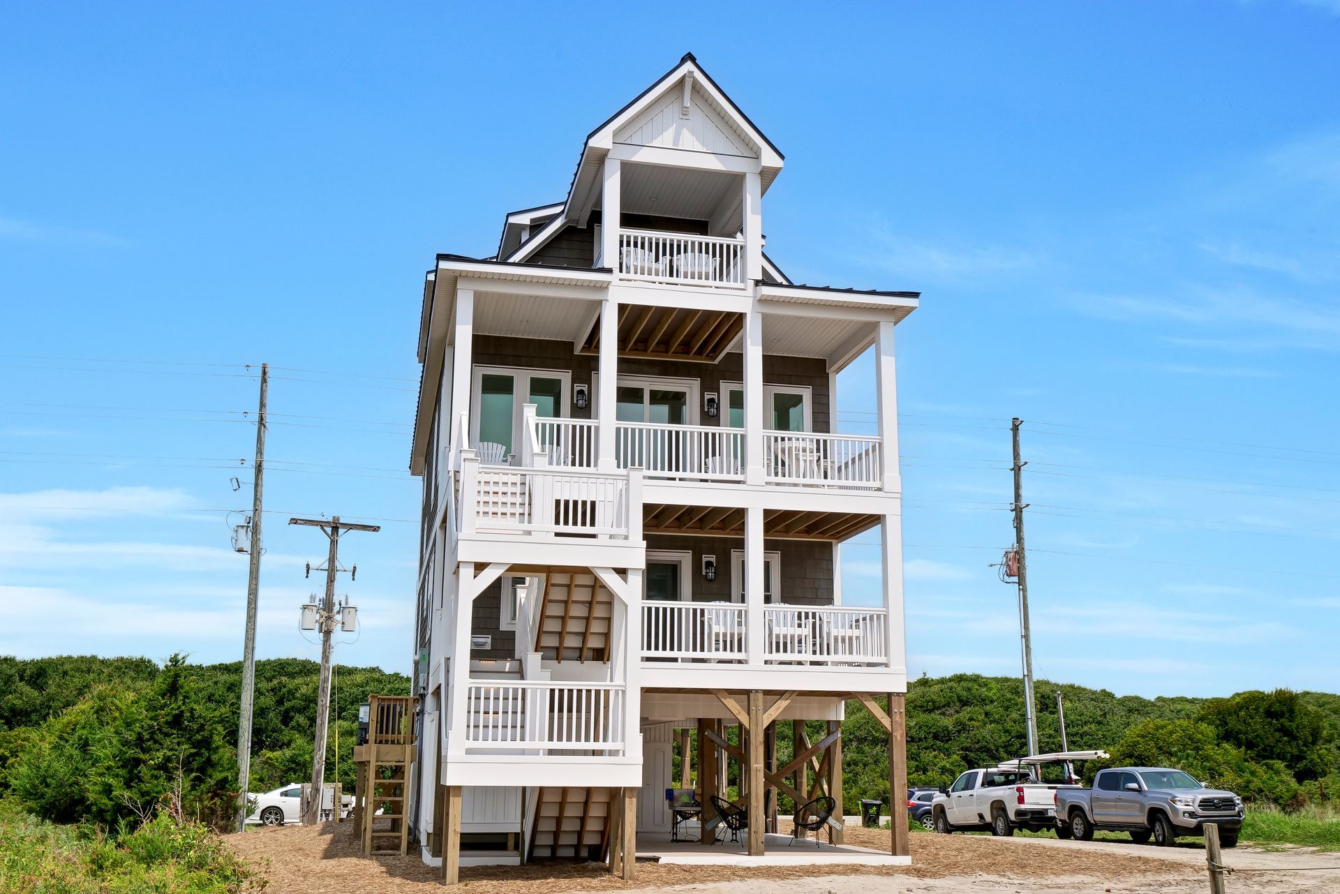 Three-story beach house on stilts with balconies. White trim, gray siding, blue sky.