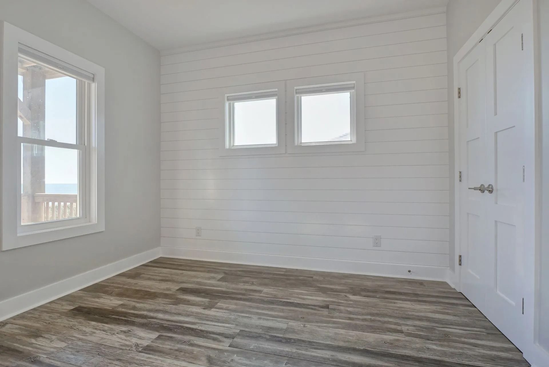 Empty bedroom with white walls, two windows, wood-look floor, and a closed door.