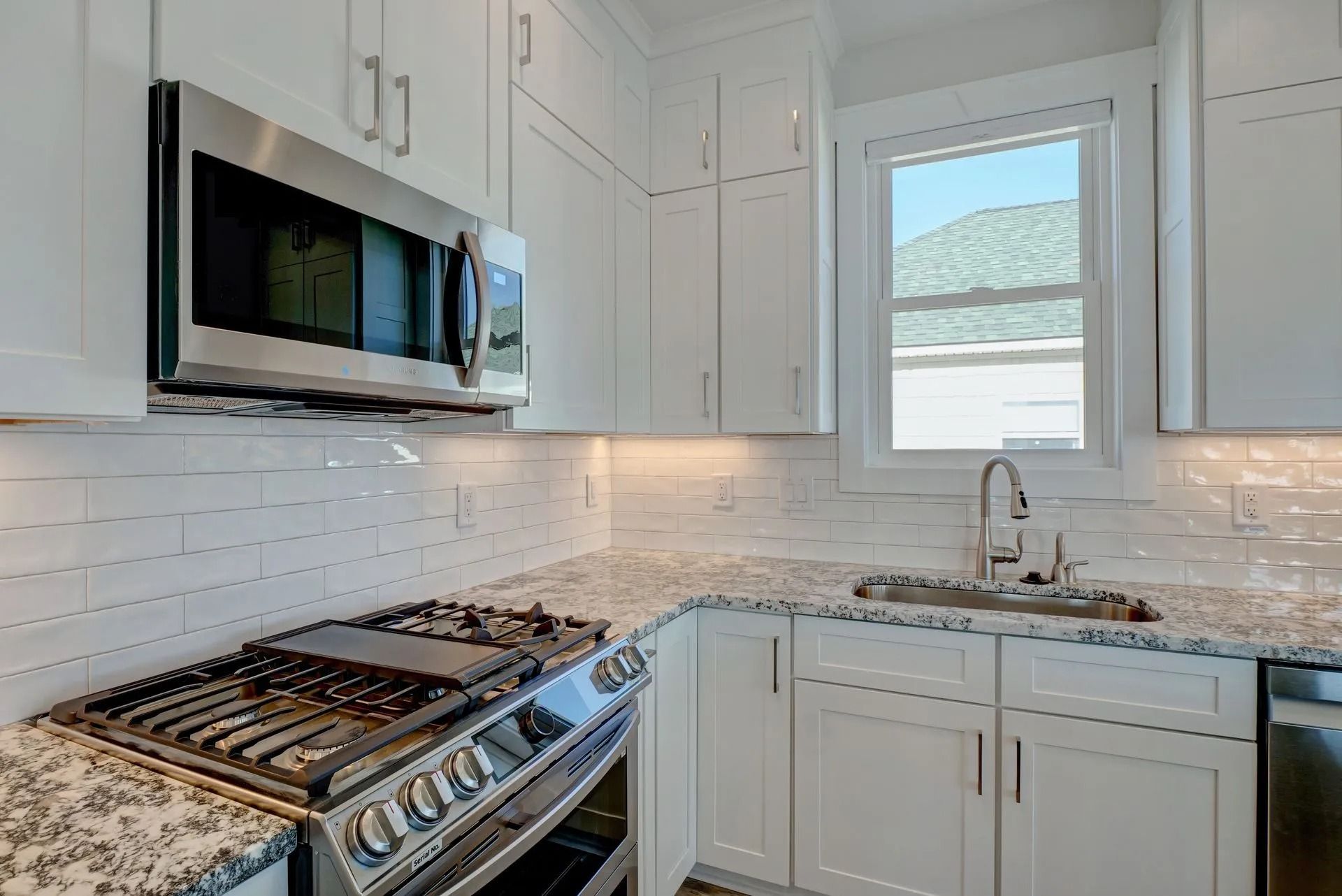 Modern white kitchen with stainless steel appliances, granite countertops, and a window.