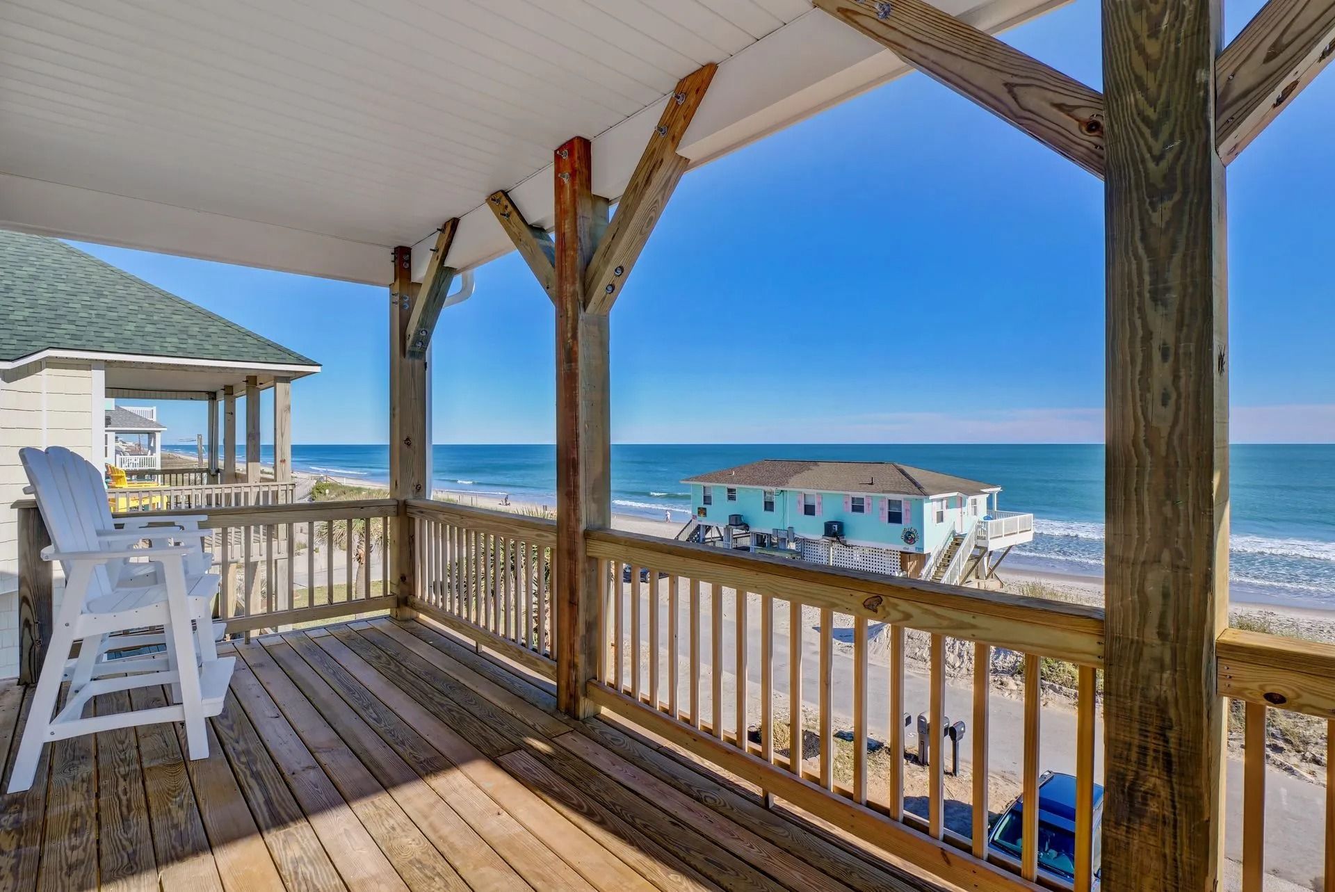 Wooden deck with a view of the ocean and beach. Two white bar stools, blue sky.