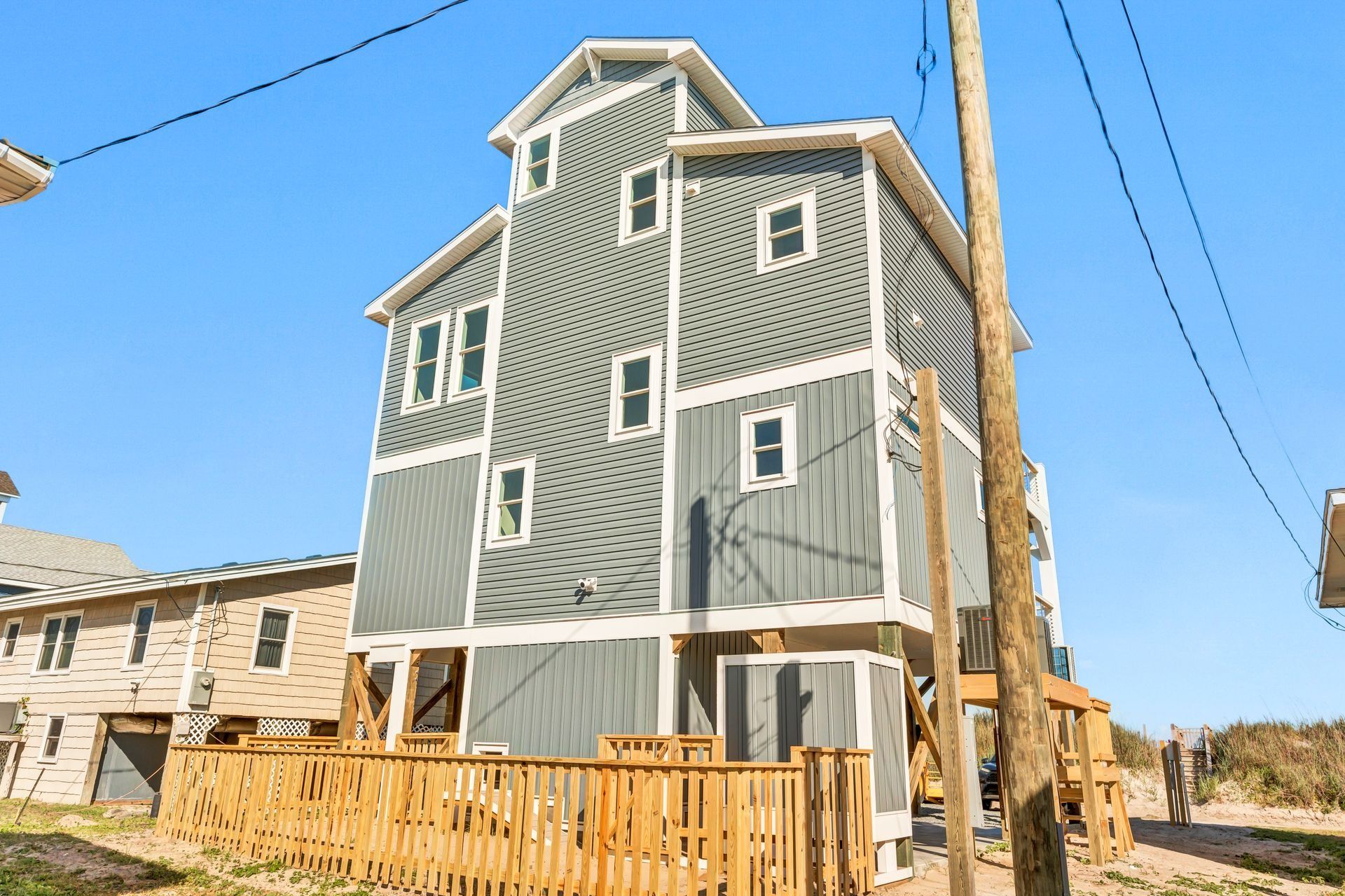 Three-story gray house with white trim and wooden deck on a beach, under a blue sky.