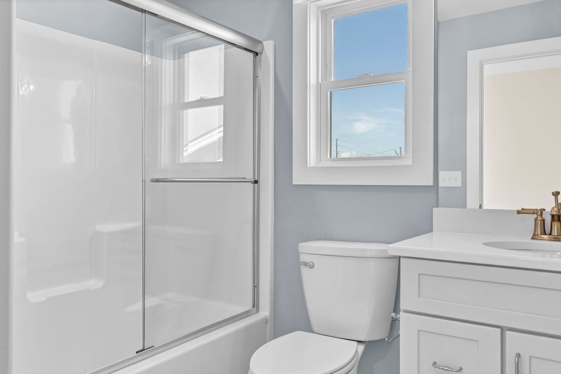 Bathroom with white fixtures, blue walls, a window showing a bright sky, and sliding glass shower doors.