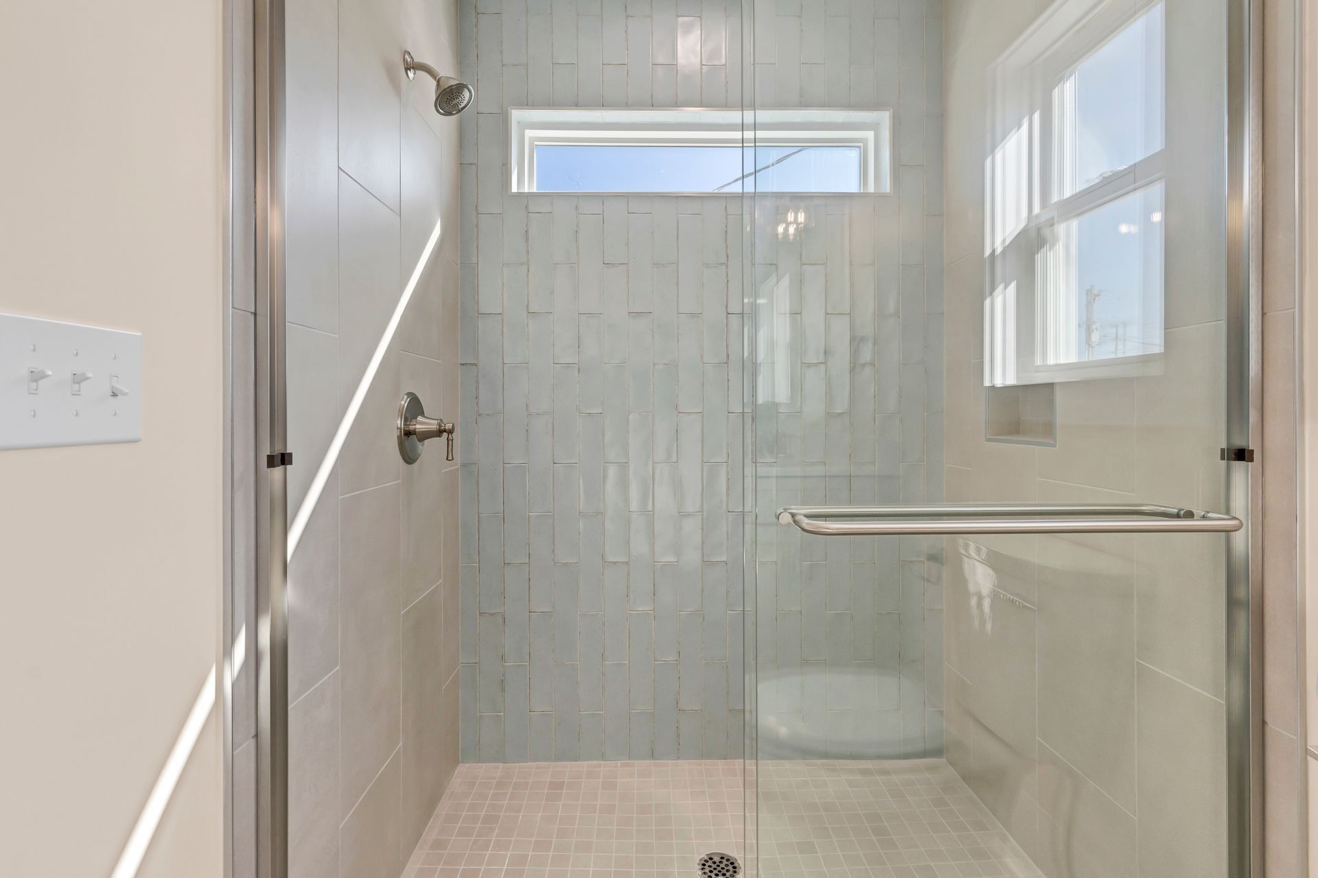 Shower stall with glass door, light blue tile, and a small window.