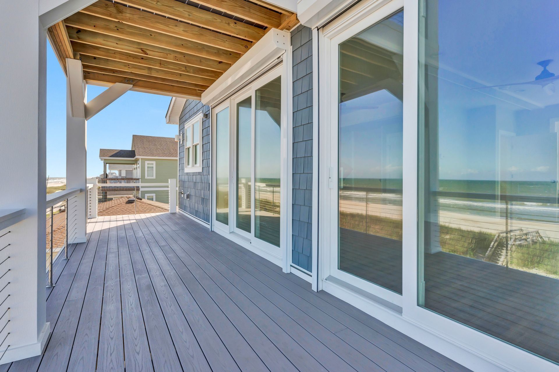 Coastal porch with gray deck, blue siding, and large sliding glass doors, overlooking beach and ocean.