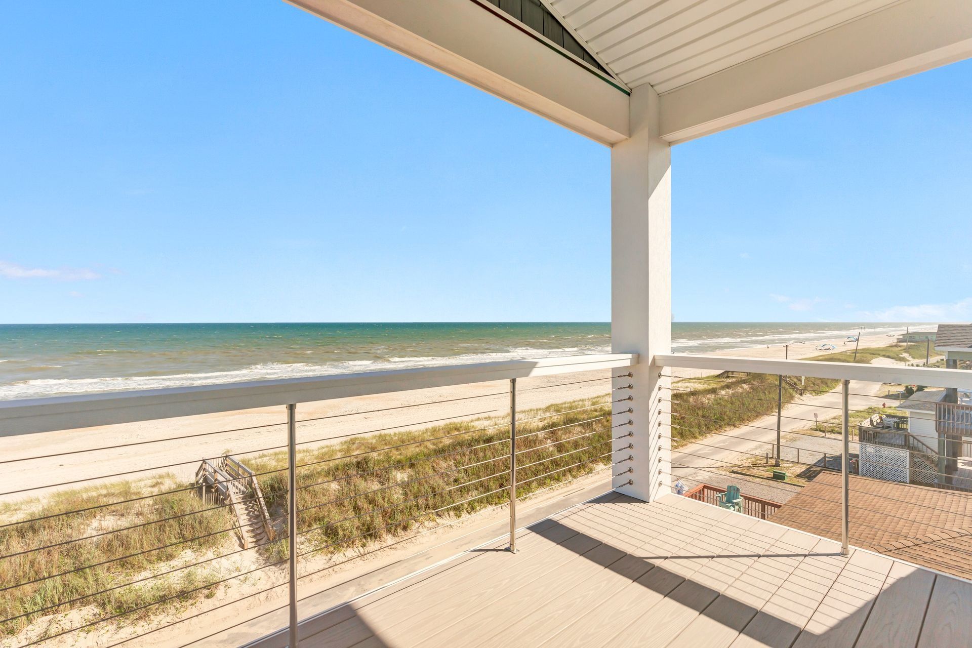 Beach view from a covered deck with glass railing. Sandy beach, ocean, and blue sky visible.