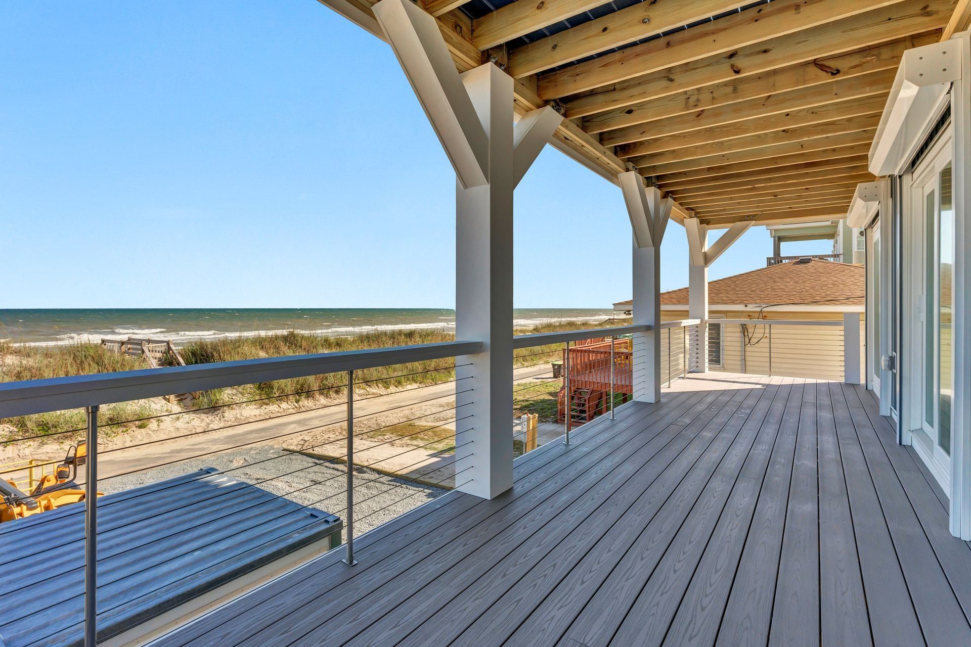Deck with a view of the beach and ocean, white pillars, gray decking, and blue sky.