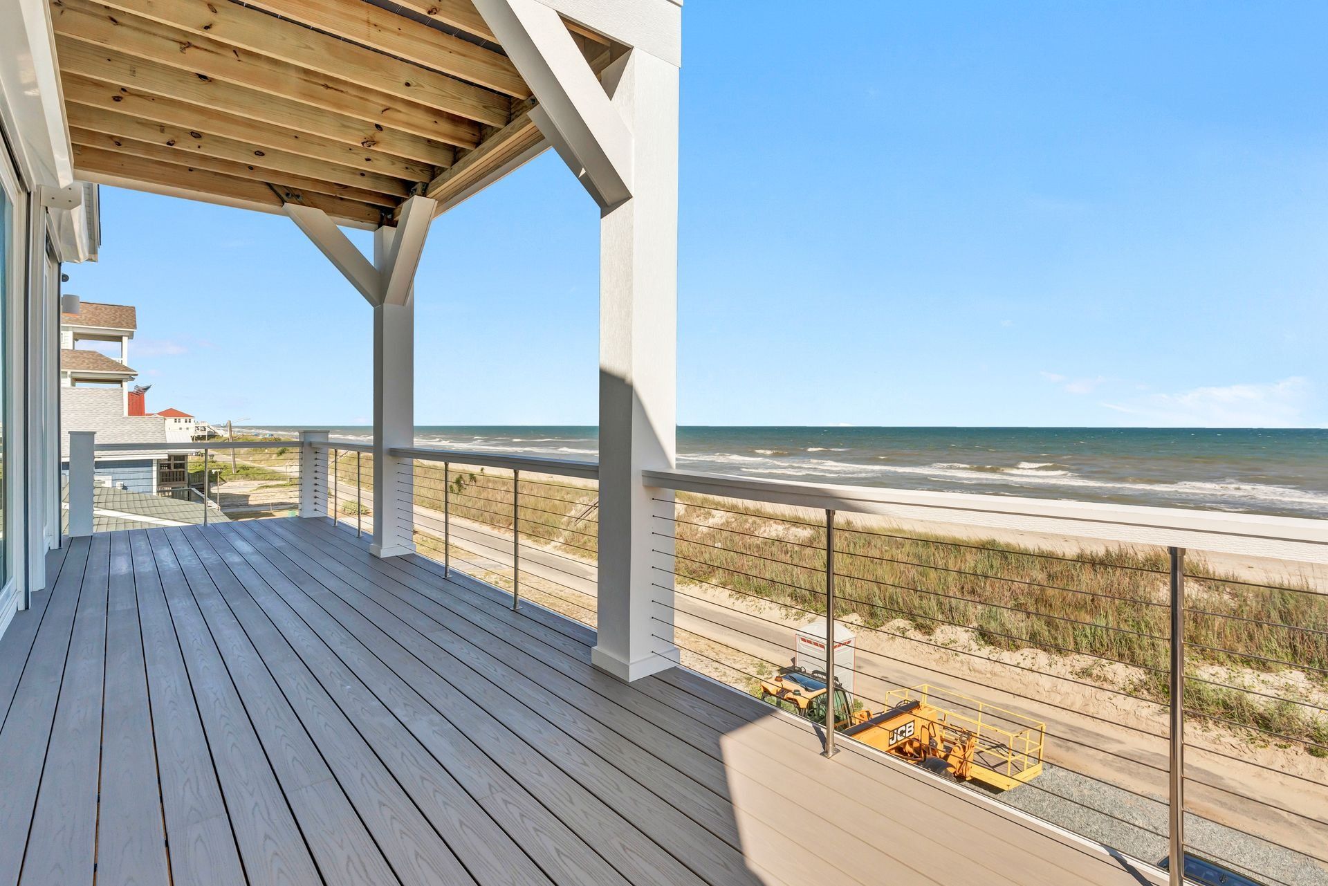 Deck overlooking the beach and ocean with blue sky. Wooden deck, white posts, and metal railings.