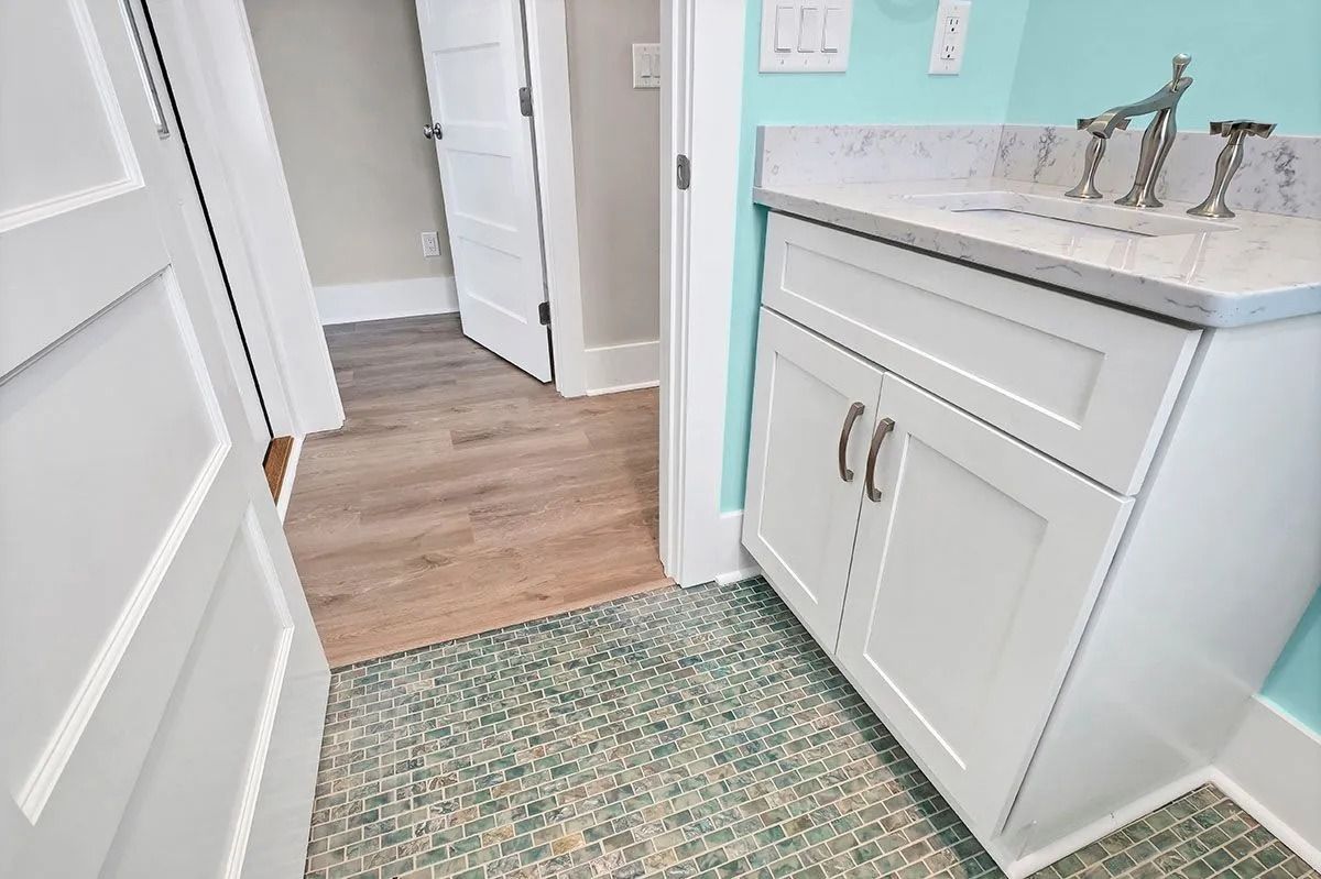 White bathroom vanity with gray countertop, teal wall, and green rug next to doorway with wood floor.