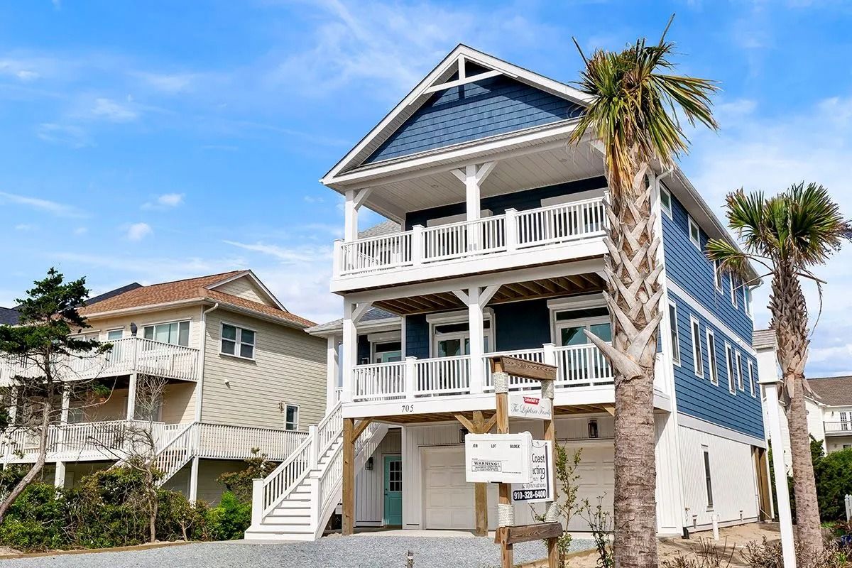 Three-story blue and white beach house with balconies, stairs, and palm trees under a blue sky.