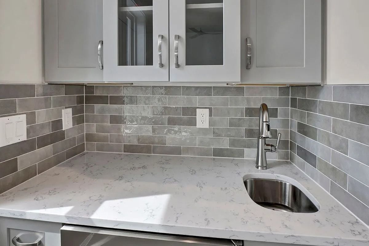 Gray tiled kitchen backsplash above a white countertop, with cabinets above and a sink.