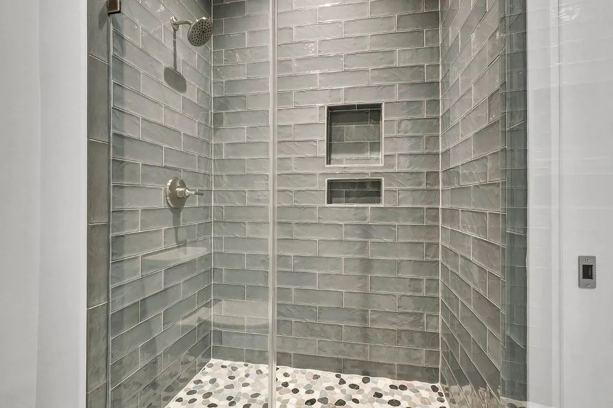 Shower with gray brick tile walls, glass door, pebble floor, and built-in shelves.