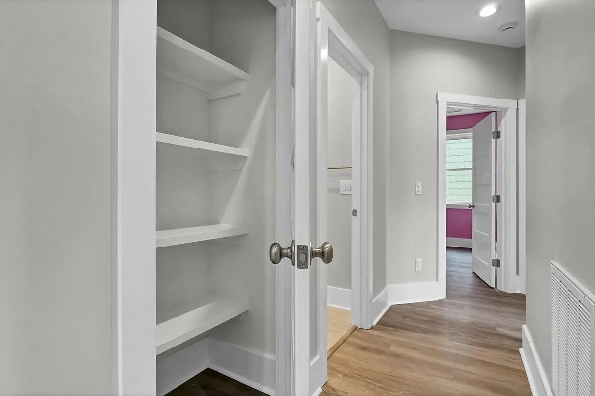 White-shelved pantry with open door, leading to a hallway with wood floor. Another door with pink wall is at the end.
