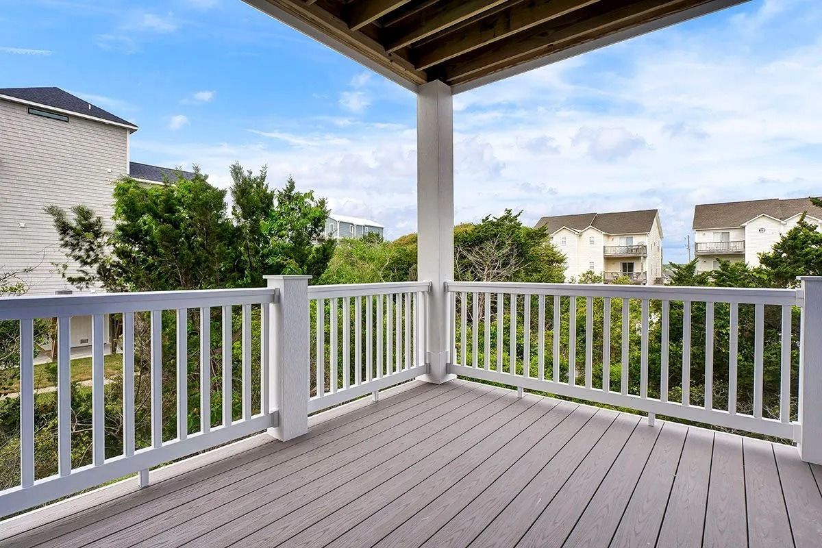 Wooden deck with white railing under a roof. Houses and trees are in the background against a blue sky.