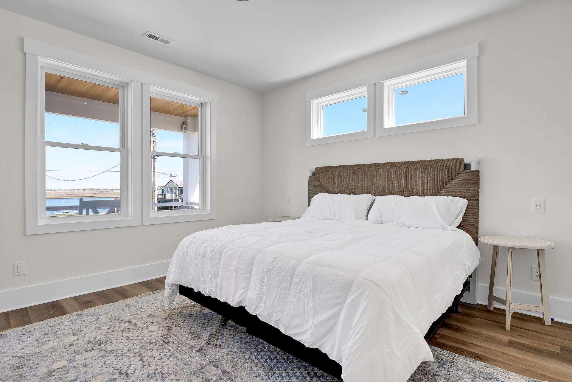 Bedroom with white bedding, headboard, and windows, light gray rug, and small white table.